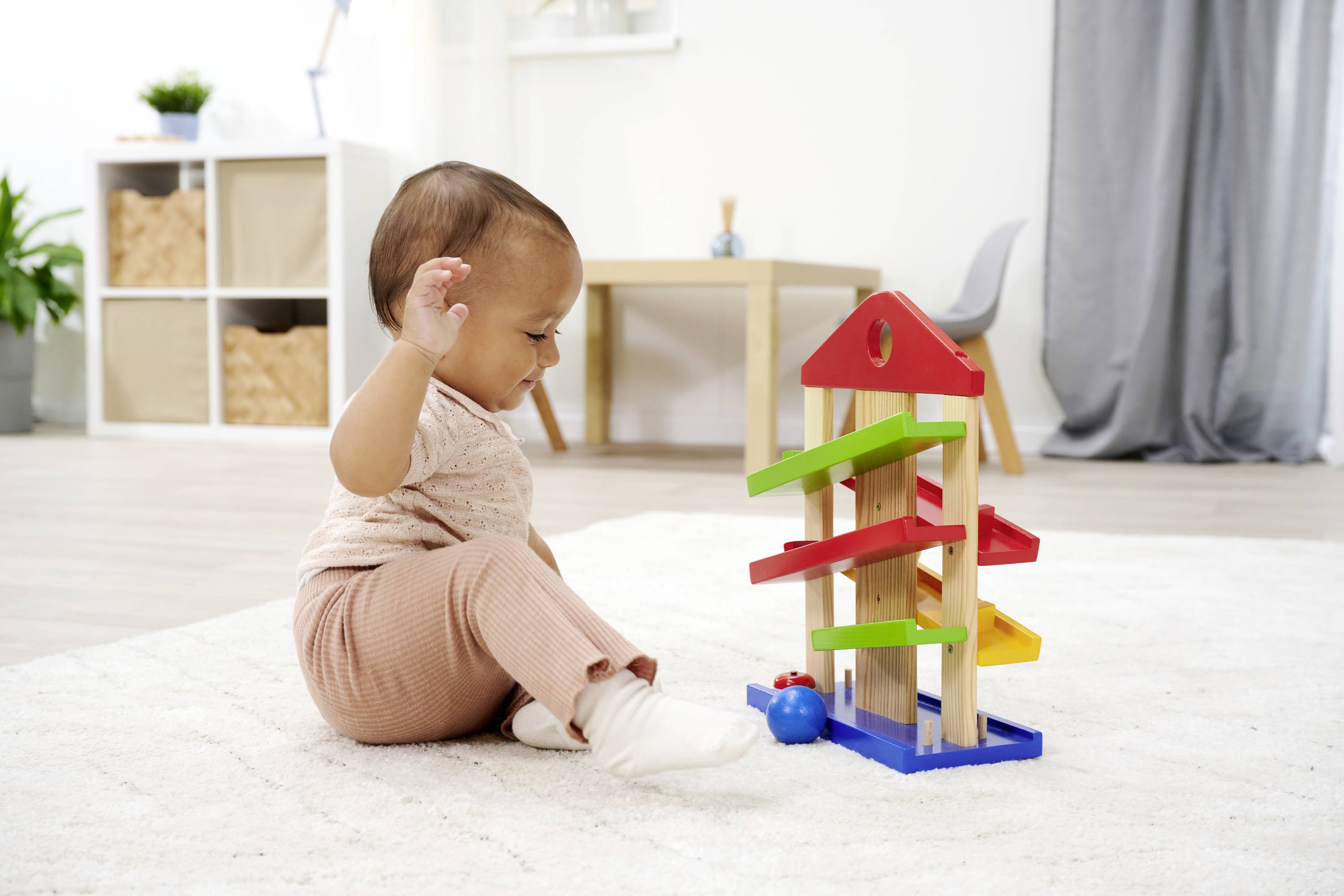 A toddler sits on a carpet, playing with a colourful wooden ball run, along whose tracks several balls are rolling.