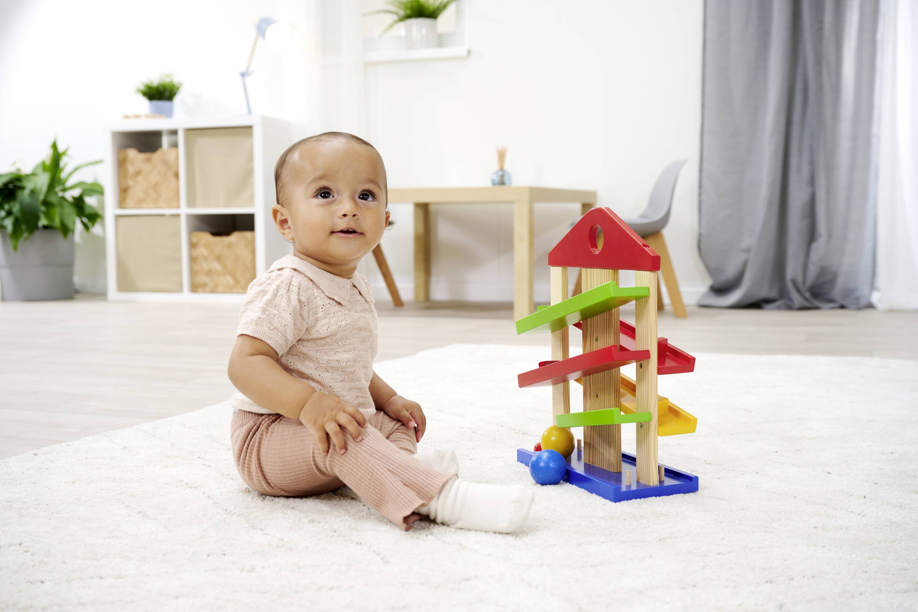 A baby is sitting on a rug and playing with a colourful marble run. Plants and furniture can be seen in the background.