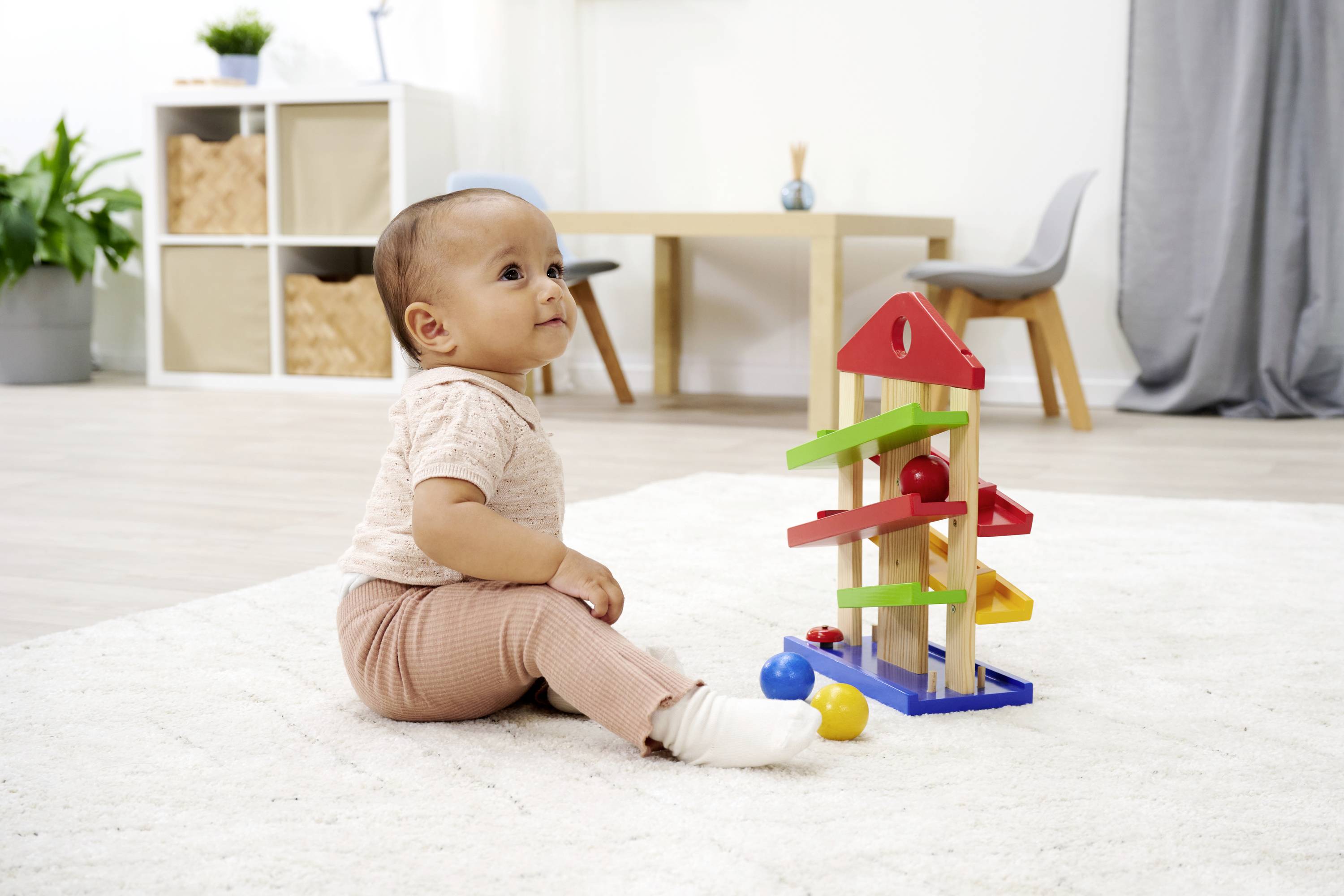 A baby is sitting on a rug and playing with a colourful wooden toy in a living room with modern furniture.