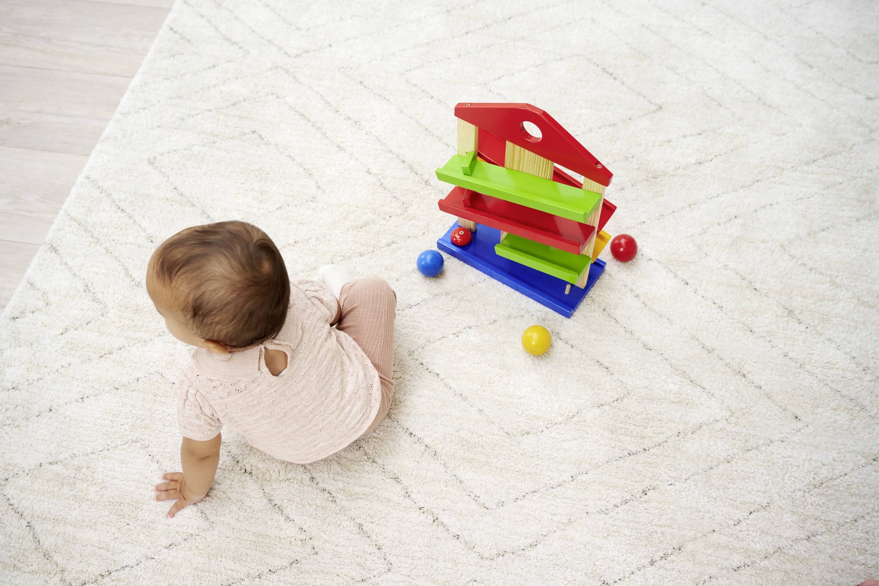 A toddler sits on a rug and plays with a colourful marble run. Three colourful balls are scattered around the marble run.
