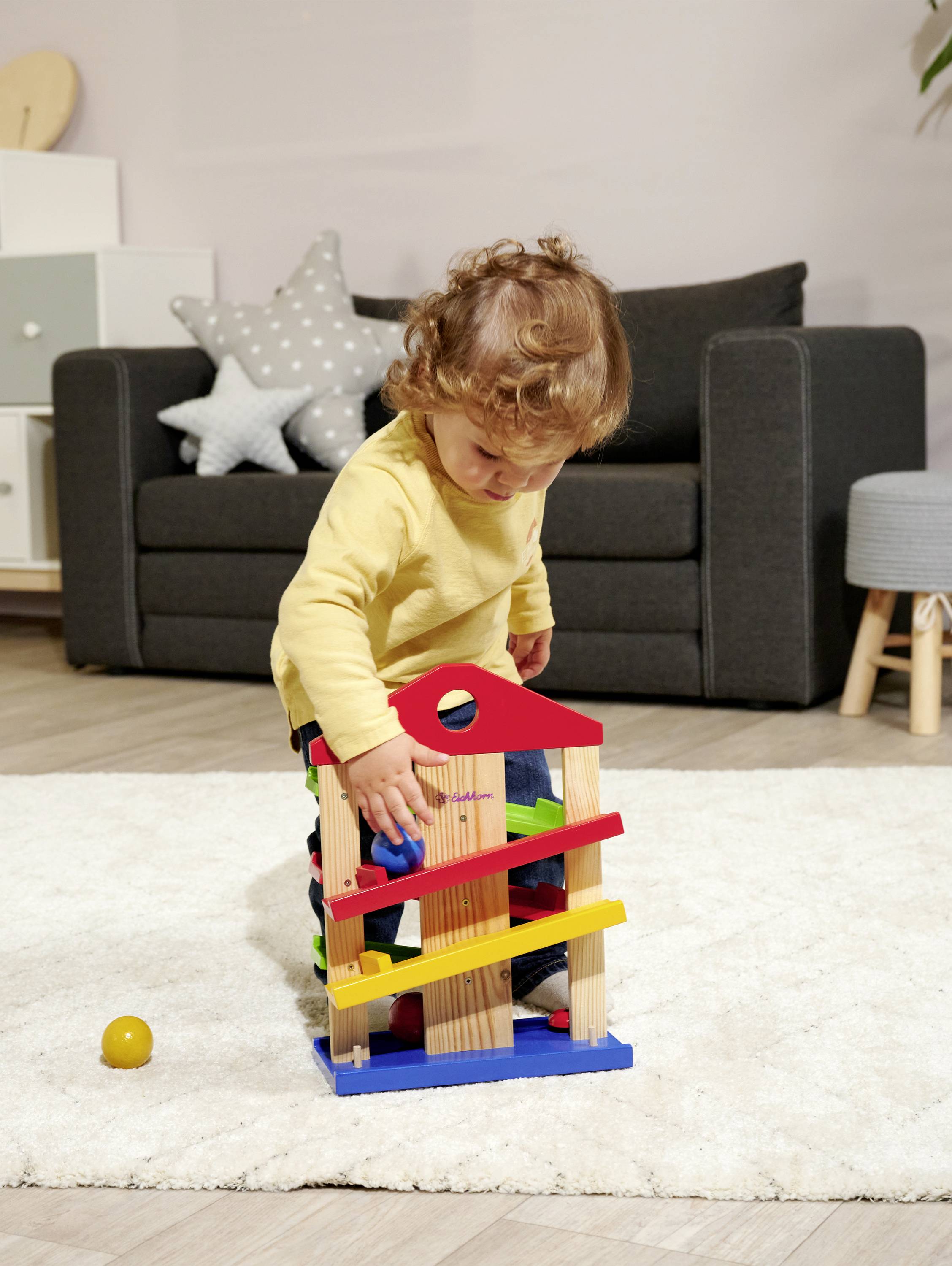 A child is playing on the carpet with a wooden marble run in the living room. In the background, there is a sofa and decorative cushions.