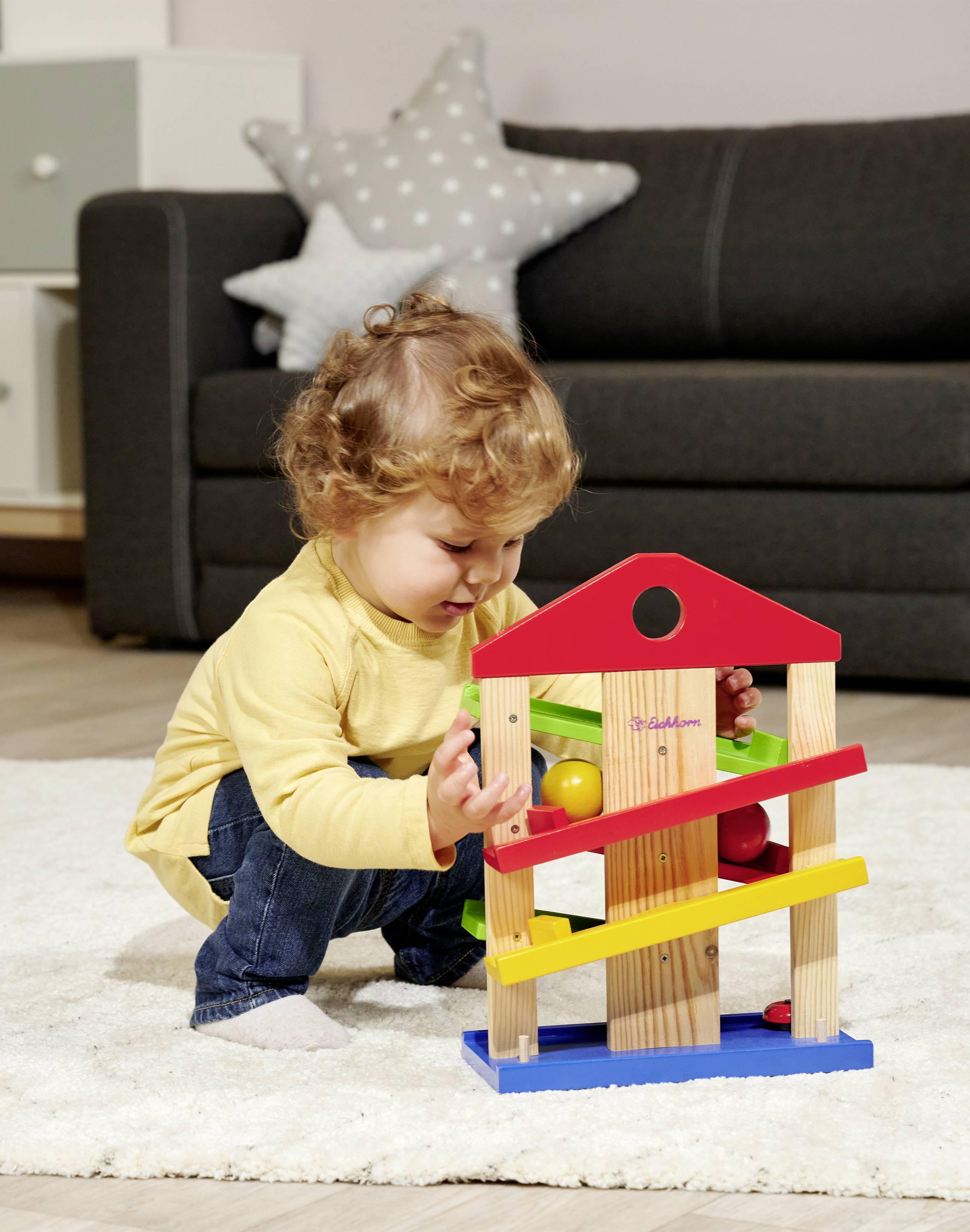 A small child is playing on a carpet with a colourful wooden toy consisting of various levels and balls. A sofa is in the background.