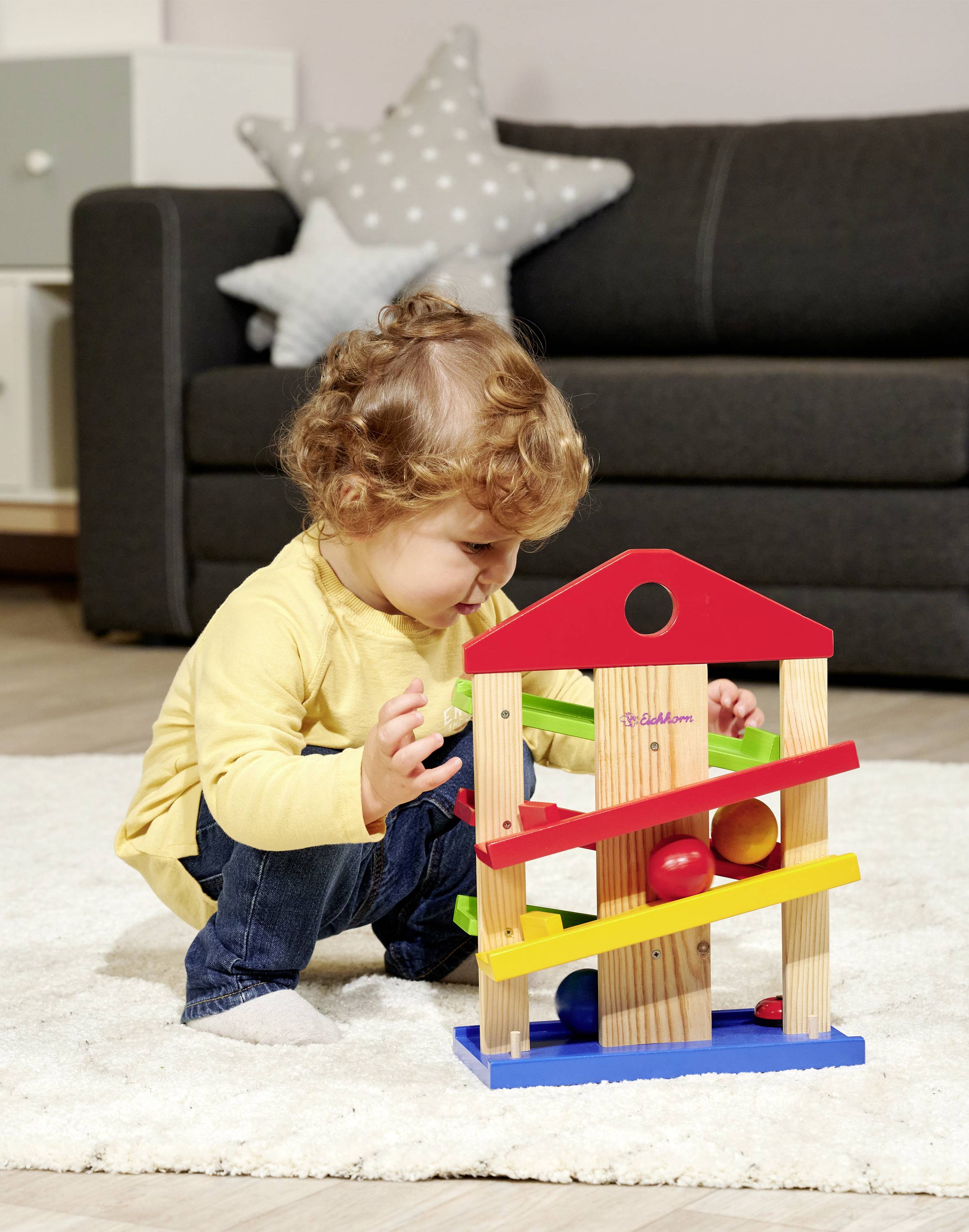 A toddler is playing on the carpet with a colourful wooden marble run, next to a grey sofa and a star-shaped cushion.