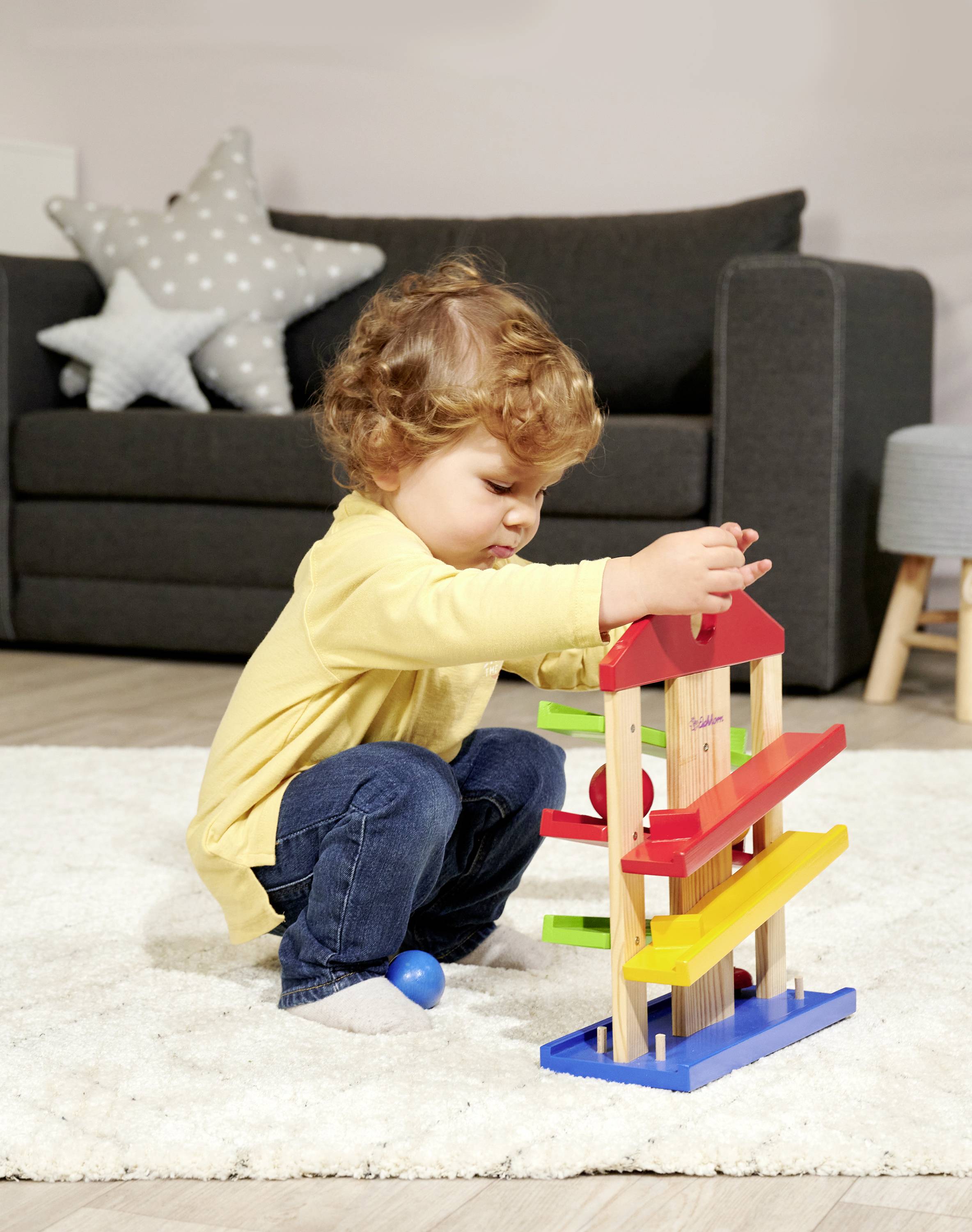 A toddler is playing with a colourful wooden toy on a carpet in the living room. In the background, there is a sofa and a footstool.