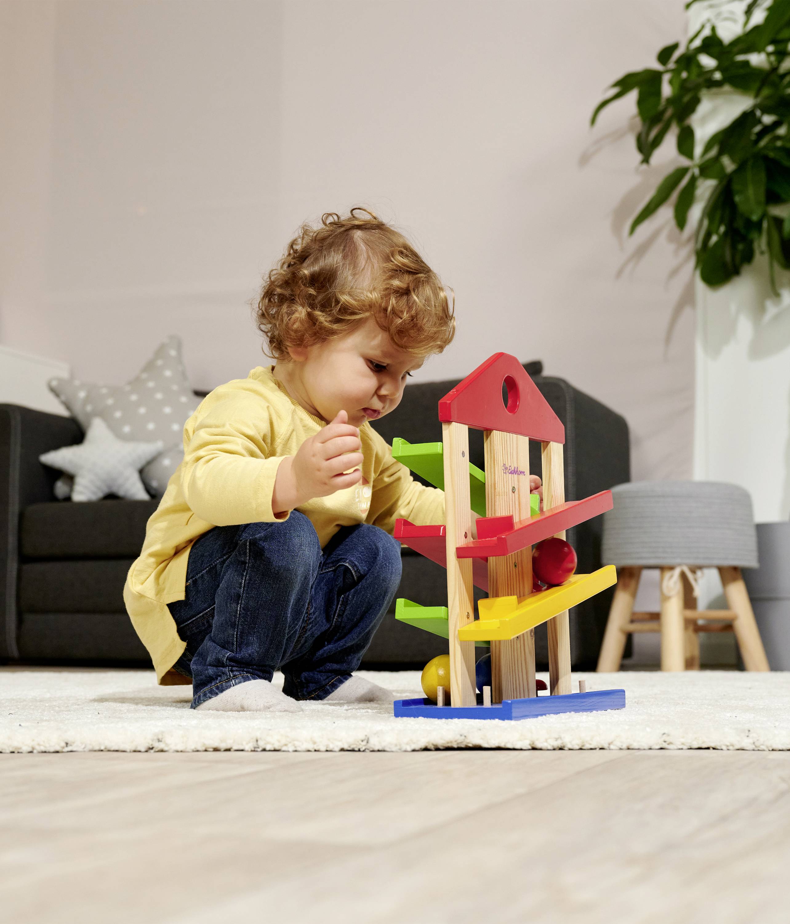 A child is playing intently with a colourful wooden toy in a living room. In the background, there is a grey chair and a plant.