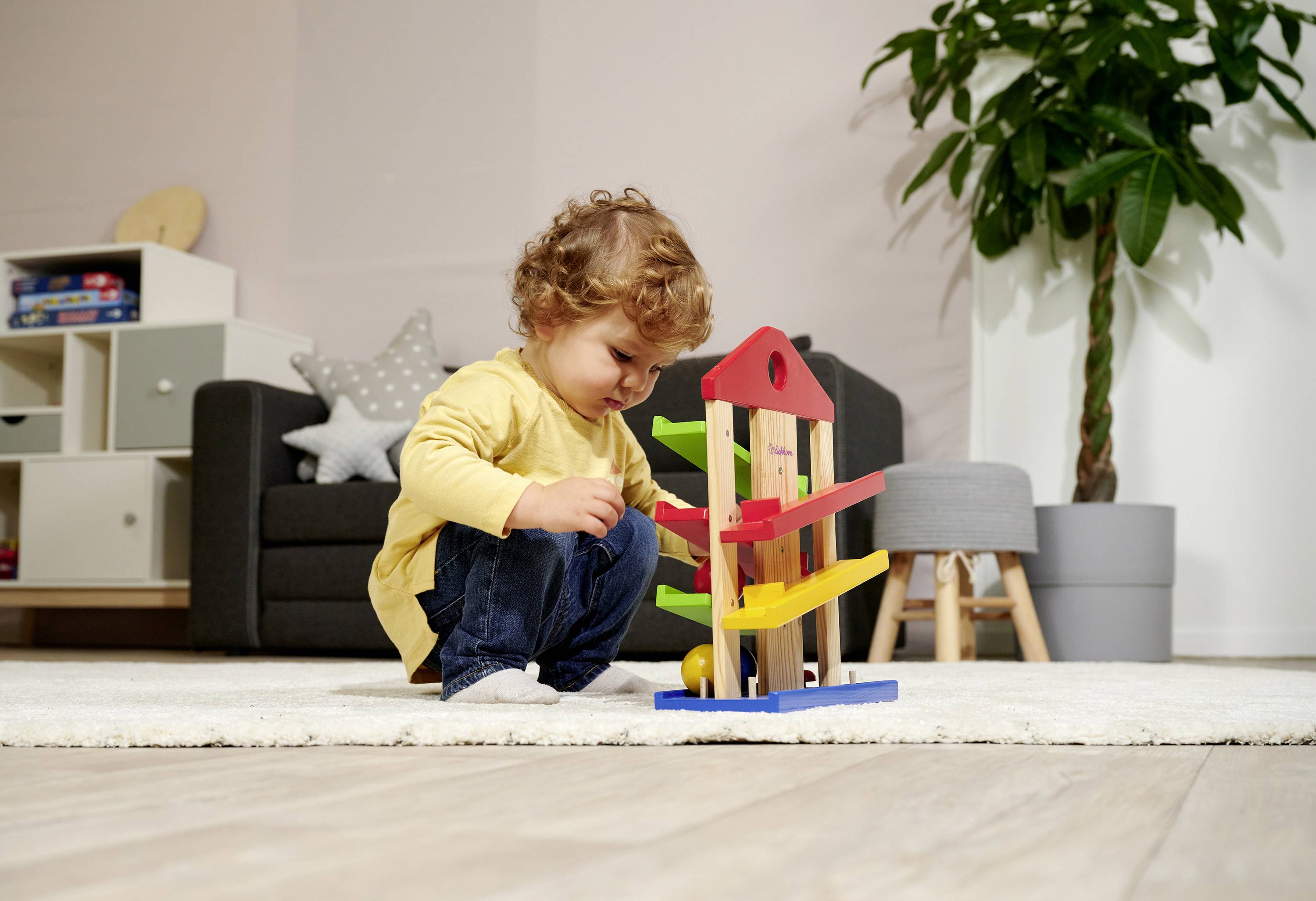 A toddler is playing with colourful wooden toys on a carpet in the living room. A sofa and a plant are visible in the background.