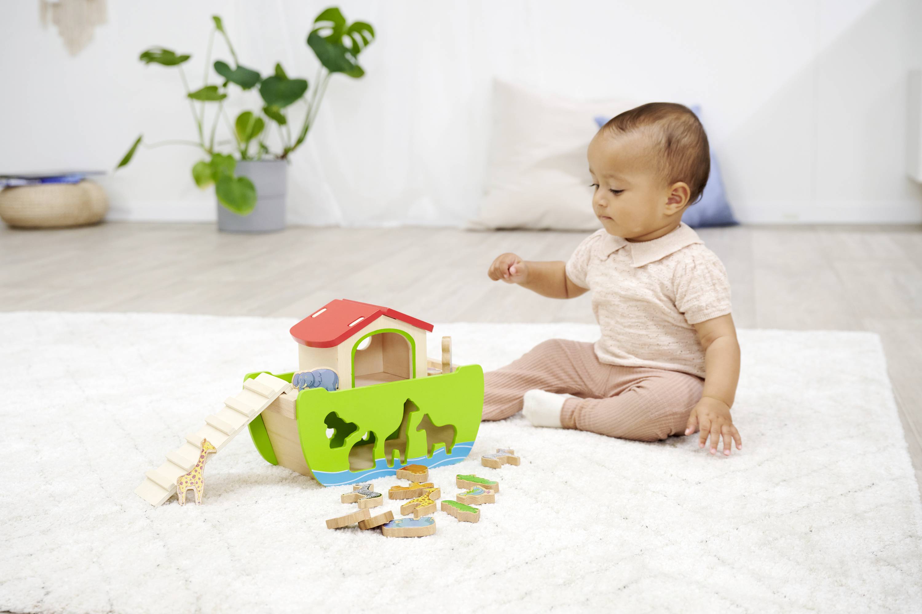 A toddler sits on a carpet, playing with a colourful wooden toy designed as an ark with animals.