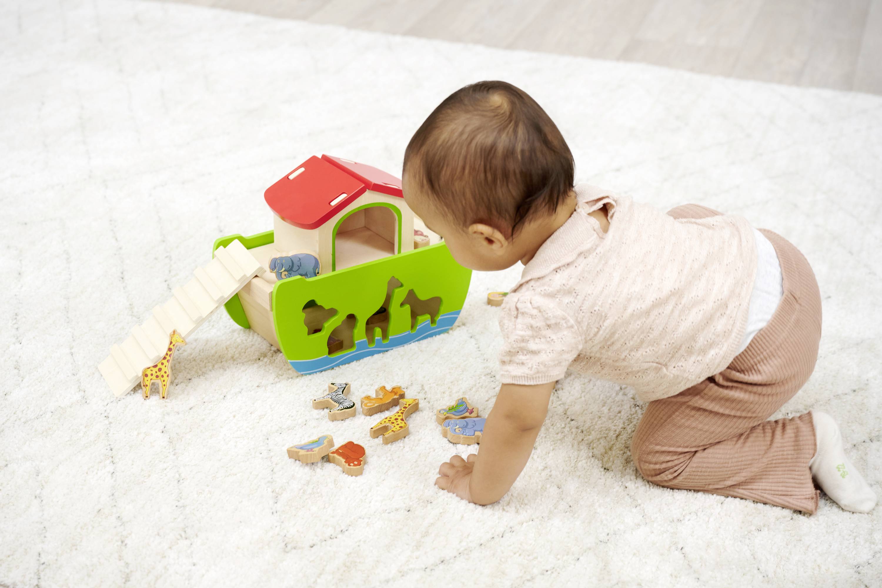 A toddler is playing on a carpet with a wooden toy ark and small animal figurines.