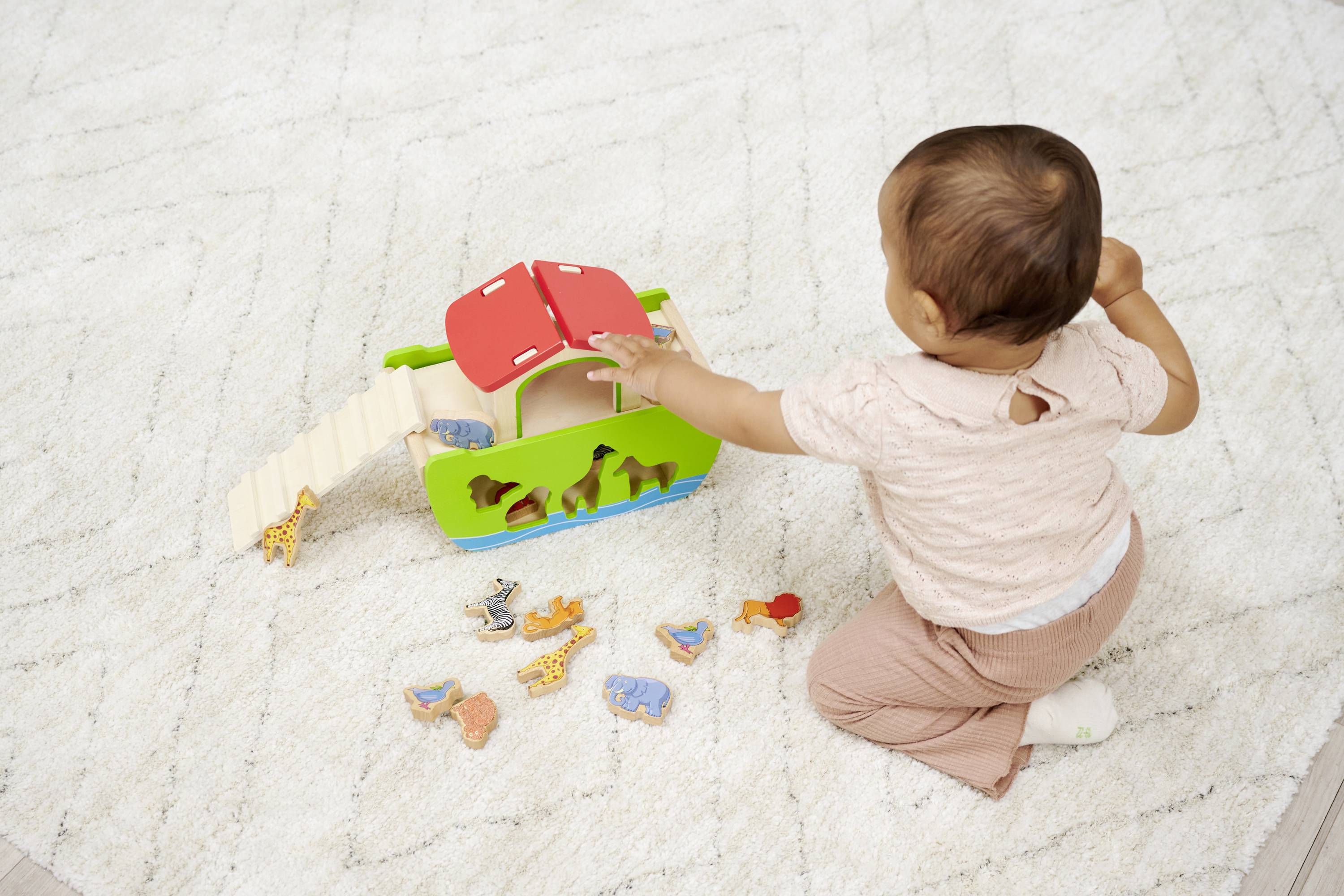 A toddler is playing with a colourful Noah's Ark toy, surrounded by small animal figurines on a carpet.