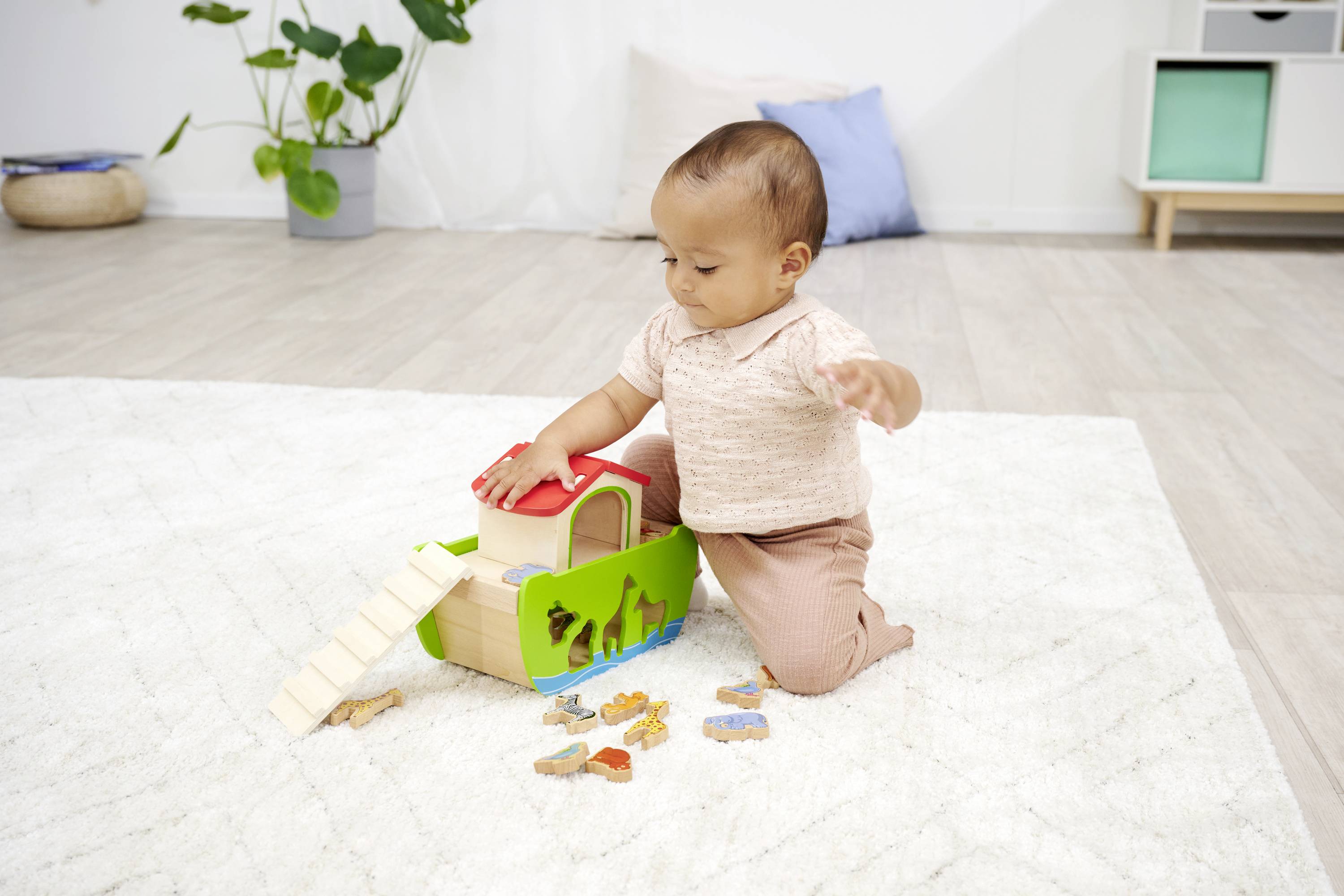 A toddler is playing on a carpet with a colourful wooden toy ark and animal figures. Plants and cushions are visible in the background.