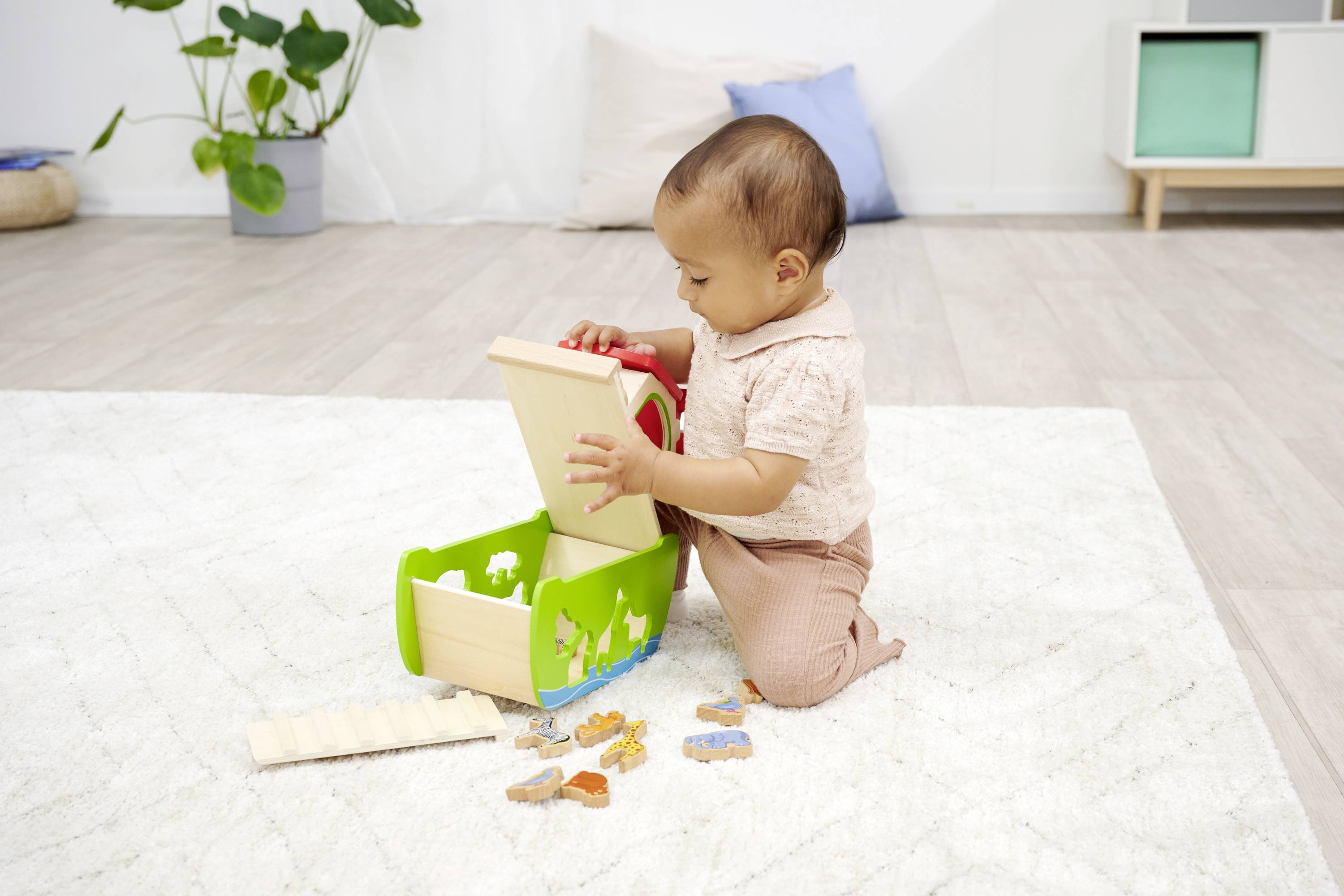 A toddler is playing on a carpet with a wooden toy box, from which they can remove shapes. Plants and furniture are visible in the background.
