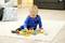 A toddler sits on a carpet and plays with colourful wooden building blocks, assembled into a train shape. A sofa and cushions are visible in the background.