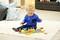 A toddler is playing on a cream-coloured carpet with a colourful wooden toy train, surrounded by cushions, in a cosy living room.