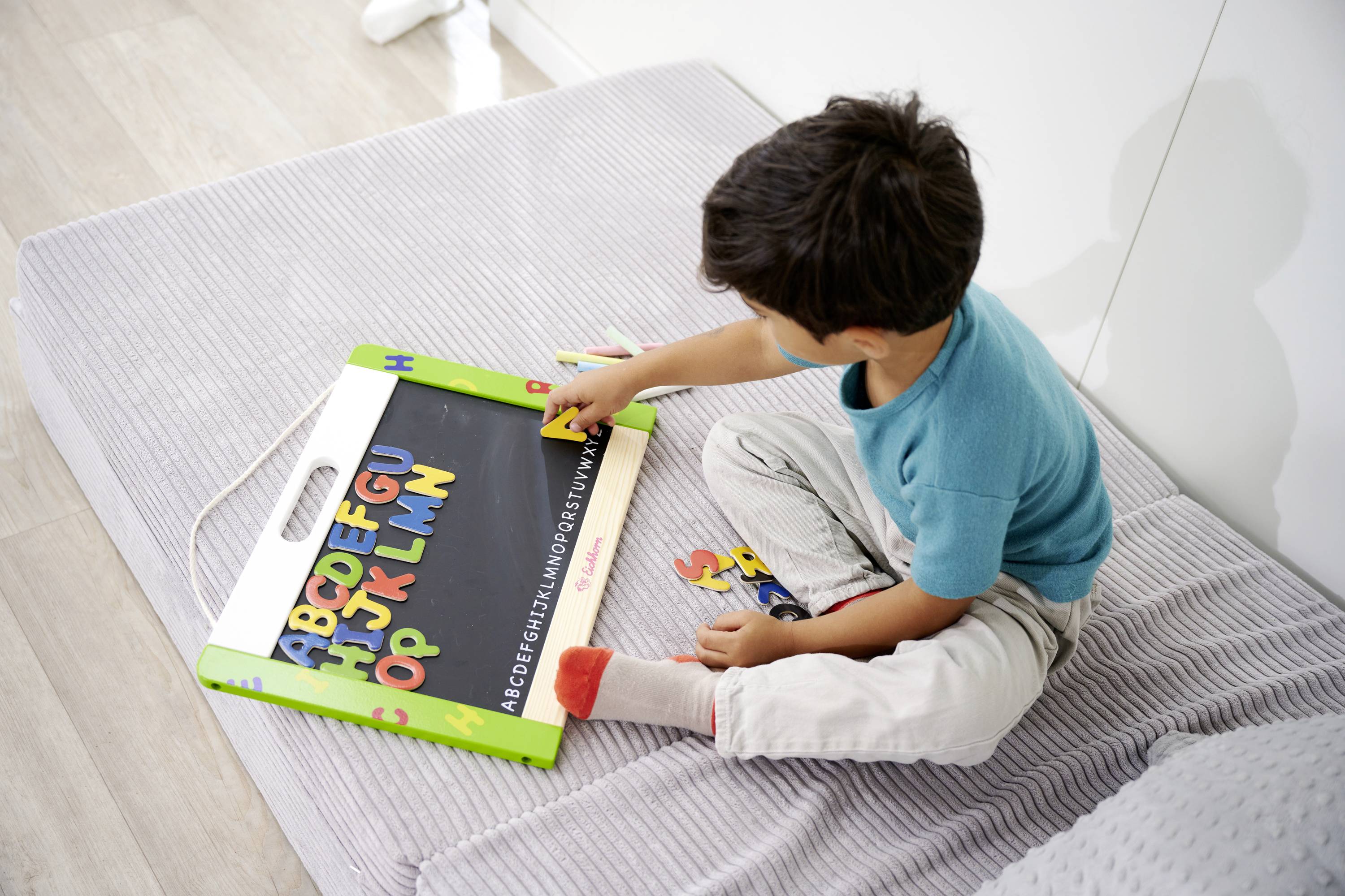 A child sits on a grey mattress and plays with a colourful alphabet game on a blackboard.