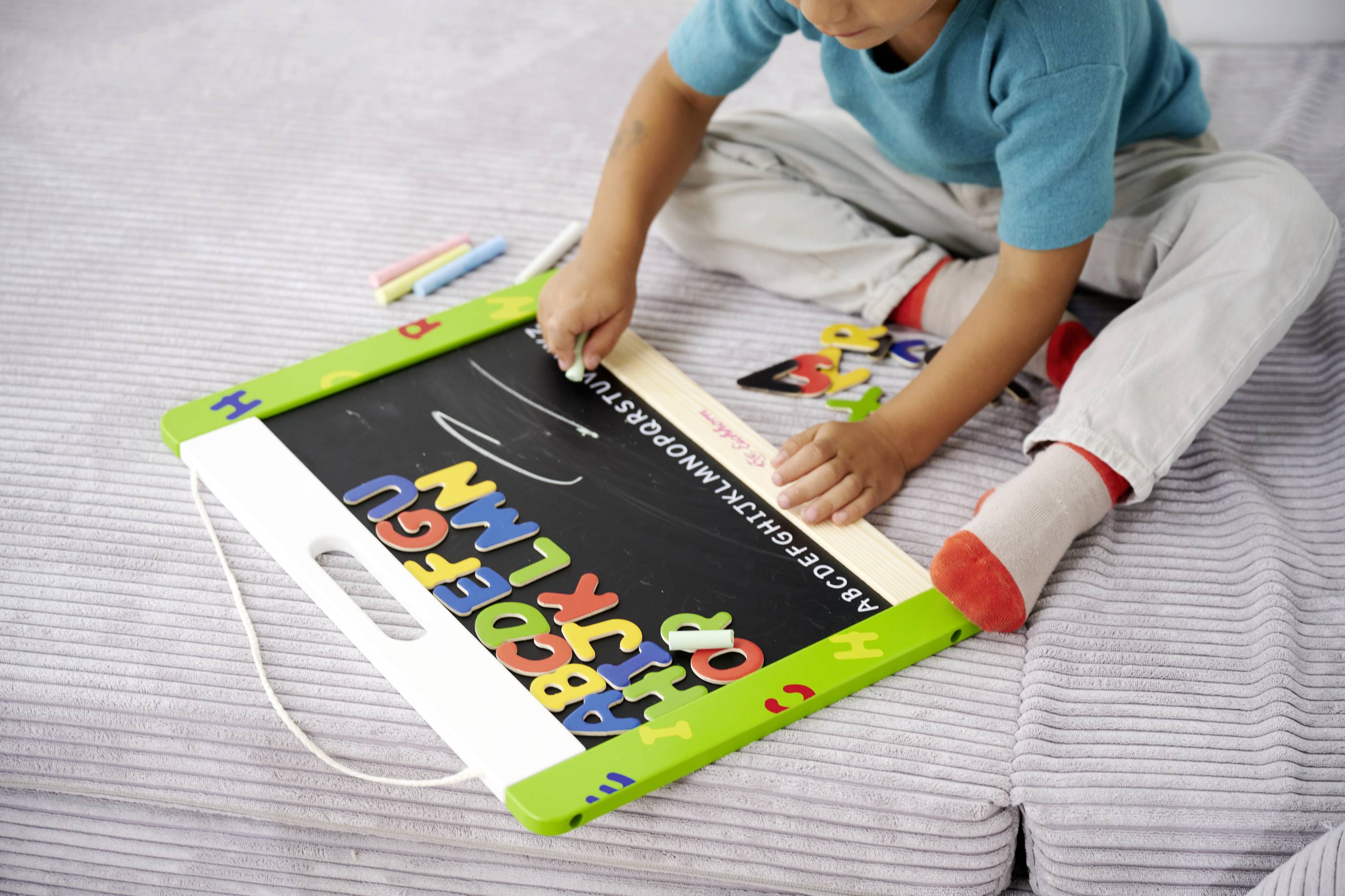 A child in blue clothing is drawing letters with chalk on a small blackboard, which is resting on a grey carpet.