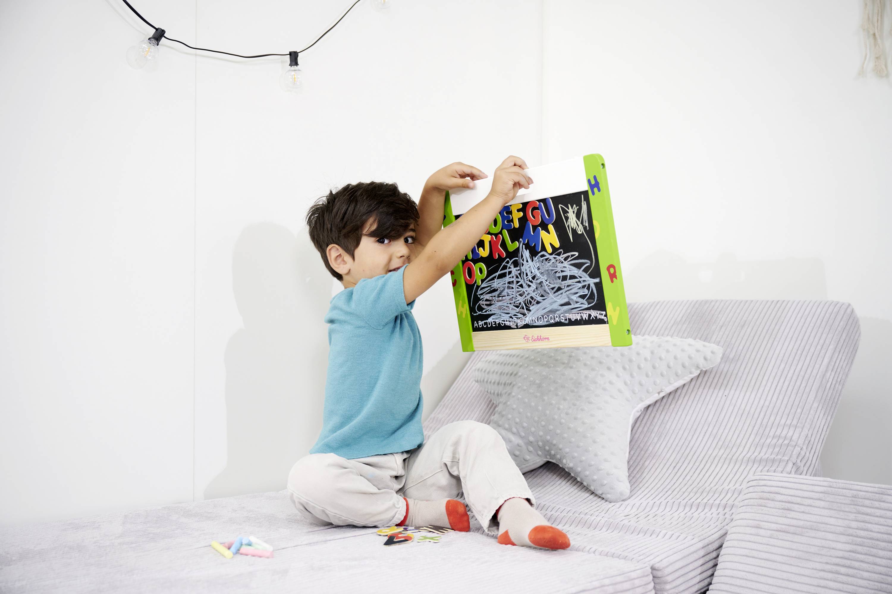 A child is sitting on a sofa, holding a small board with colourful letters and doodles.