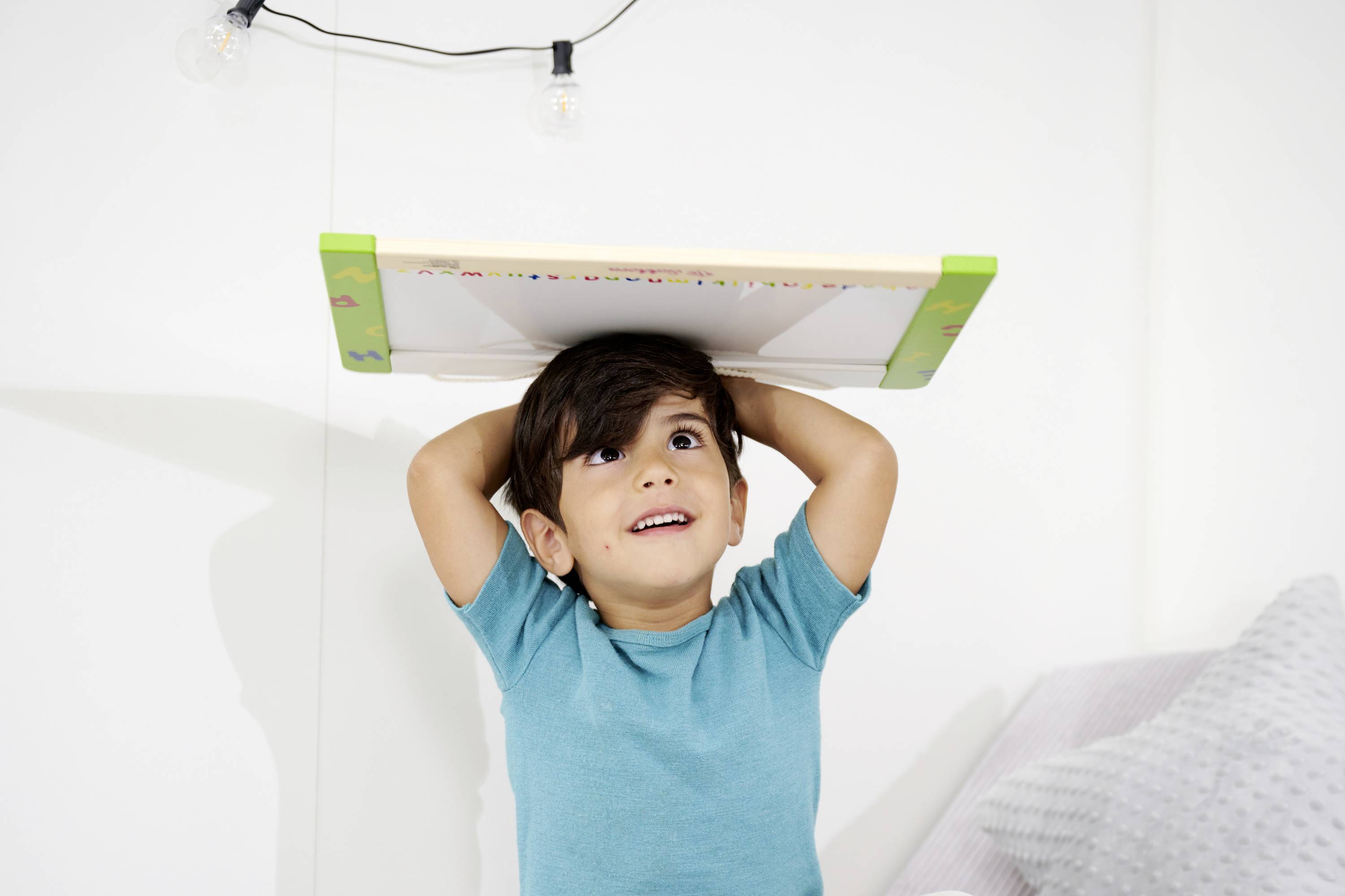 Small boy in a blue T-shirt holding a board with a colourful frame above his head and smiling. A white wall is in the background.