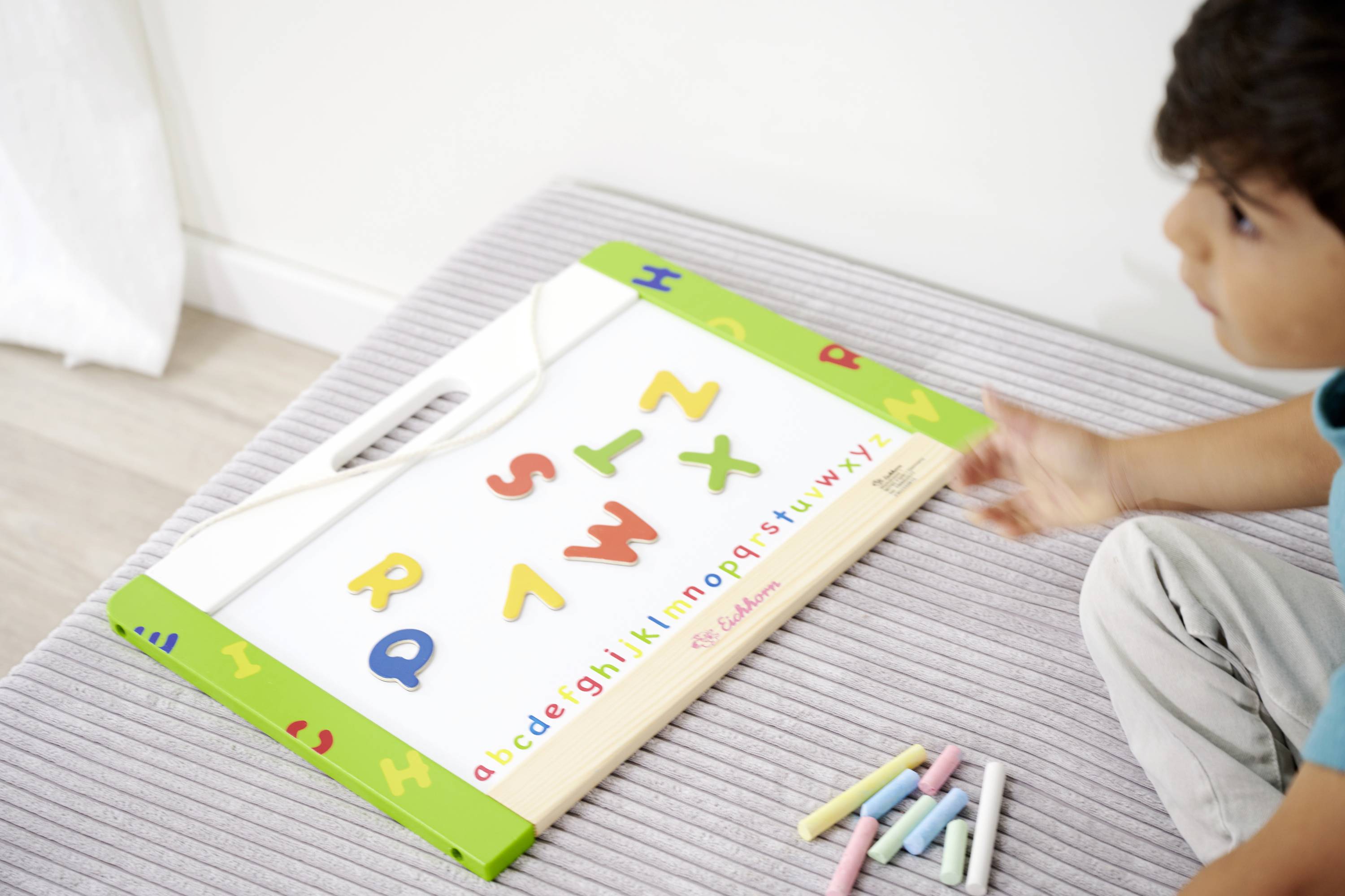 A child is sitting next to a board with colourful magnetic letters. Scattered on the side are pieces of coloured chalk.