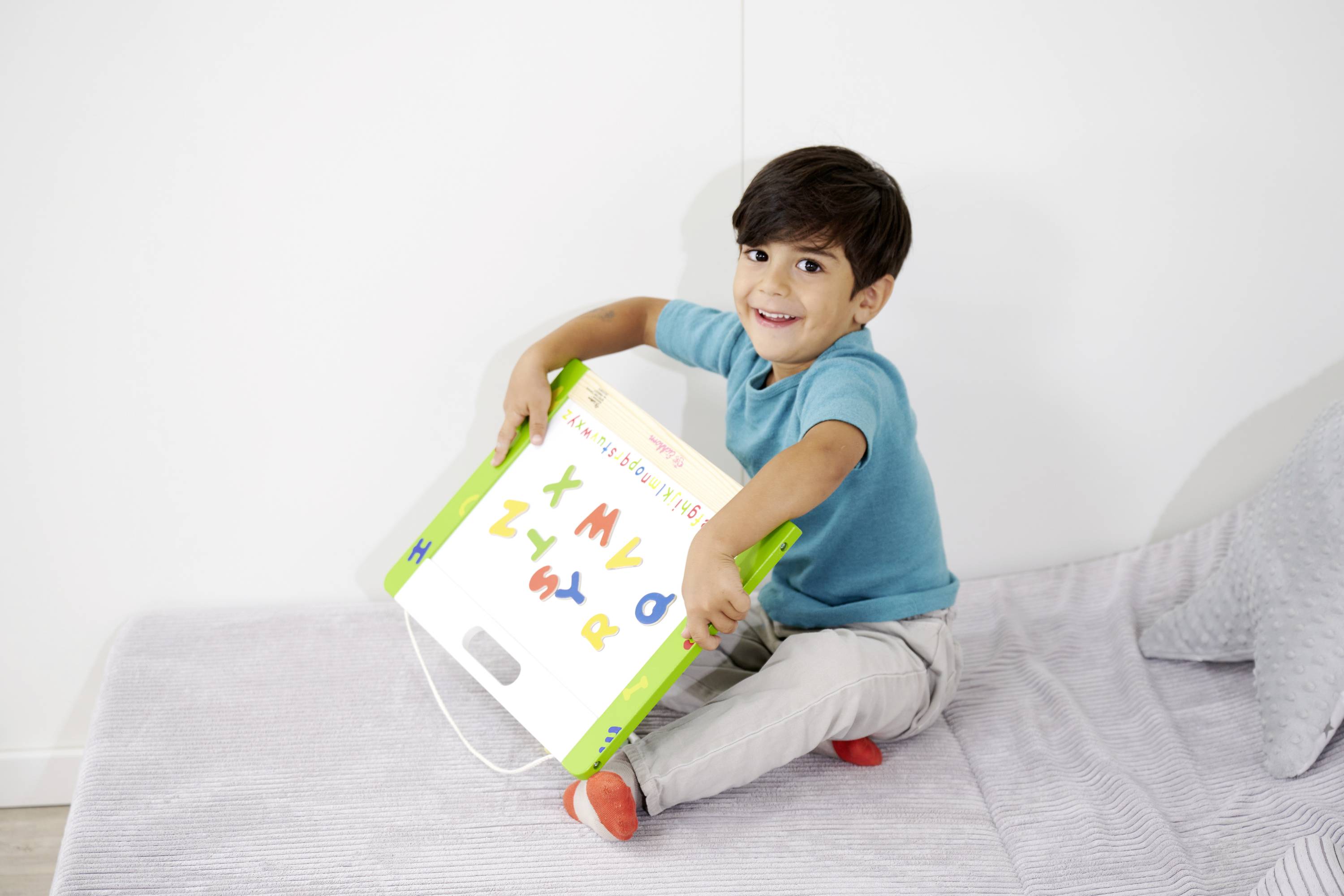 A smiling child in a blue t-shirt sits on a bed, holding a toy with colourful letters in their hands.