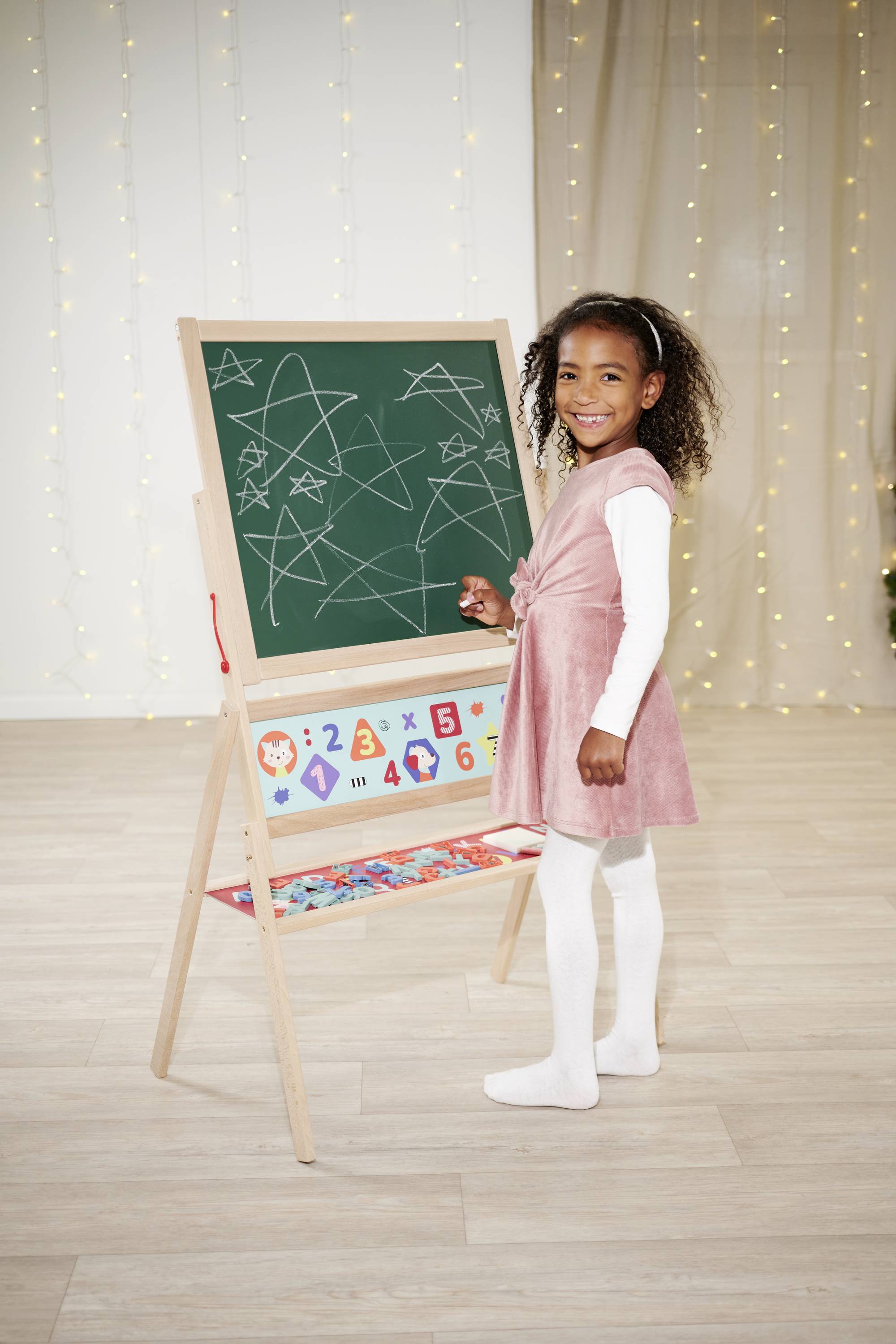 A girl stands smiling in front of a blackboard with painted stars and numbers. She is wearing a pink dress and white tights.