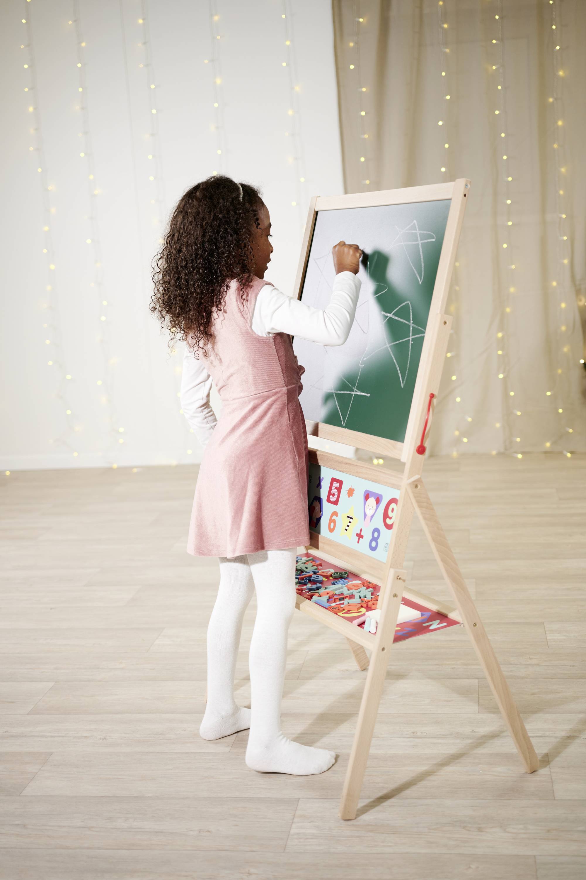 A smiling girl stands in front of a children's easel with a board full of chalk drawings in a festively decorated room.
