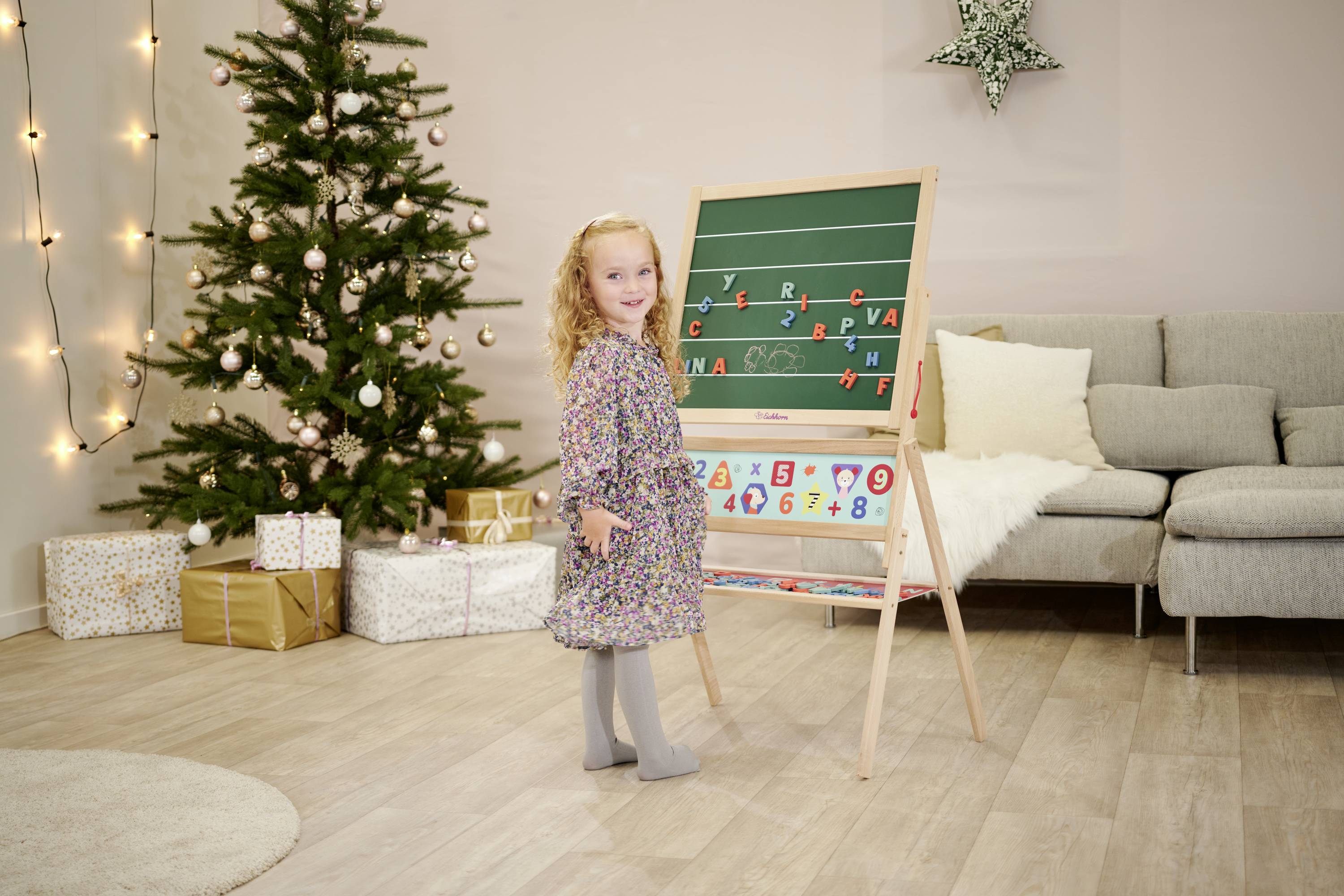 A small girl stands smiling beside a blackboard with letters and numbers visible. In the background, a decorated Christmas tree stands.