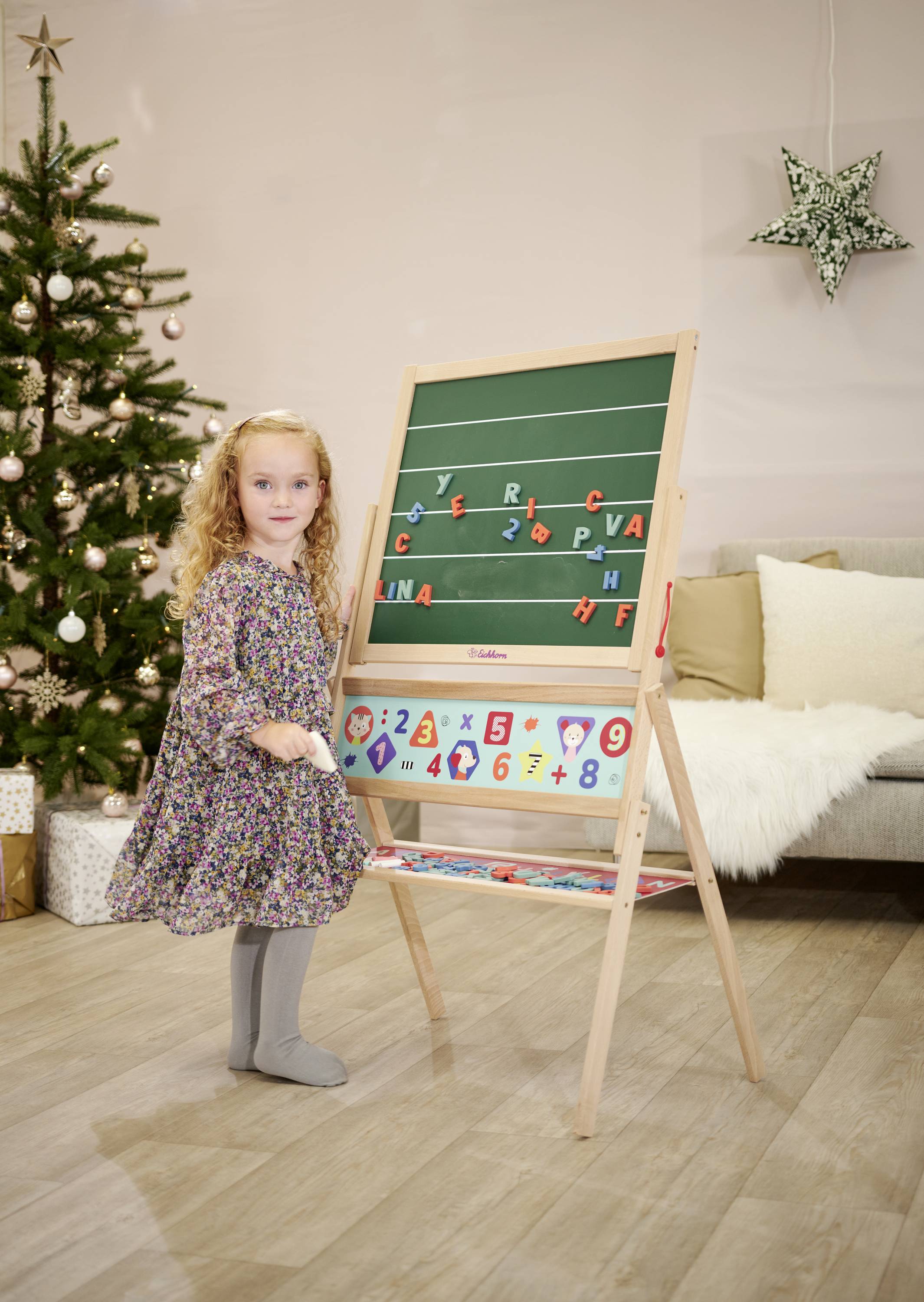 A girl is standing in front of a blackboard with letters and numbers, next to a decorated Christmas tree in the living room.