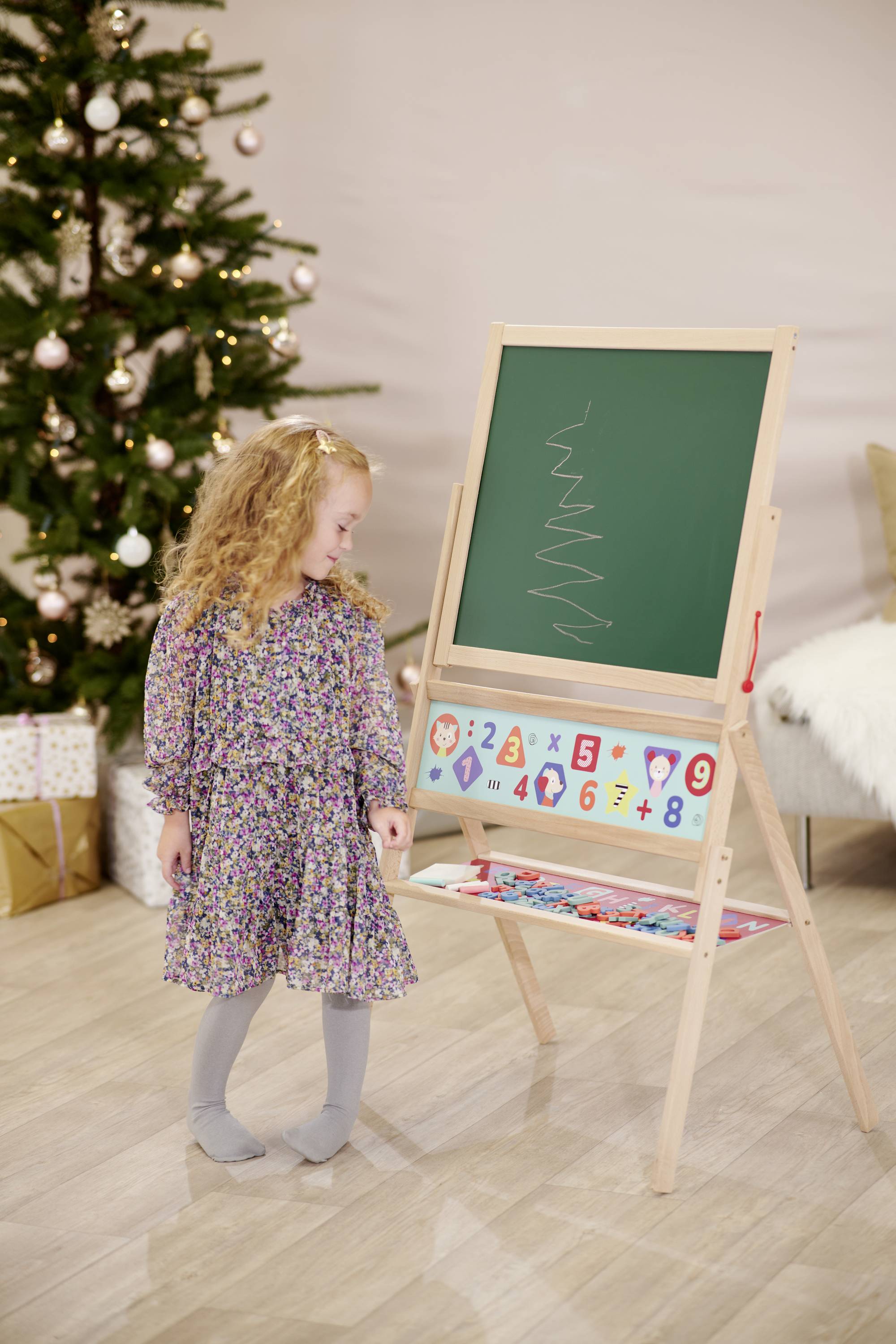 A child is standing next to a small blackboard with scribbles. In the background, a decorated Christmas tree with presents can be seen.