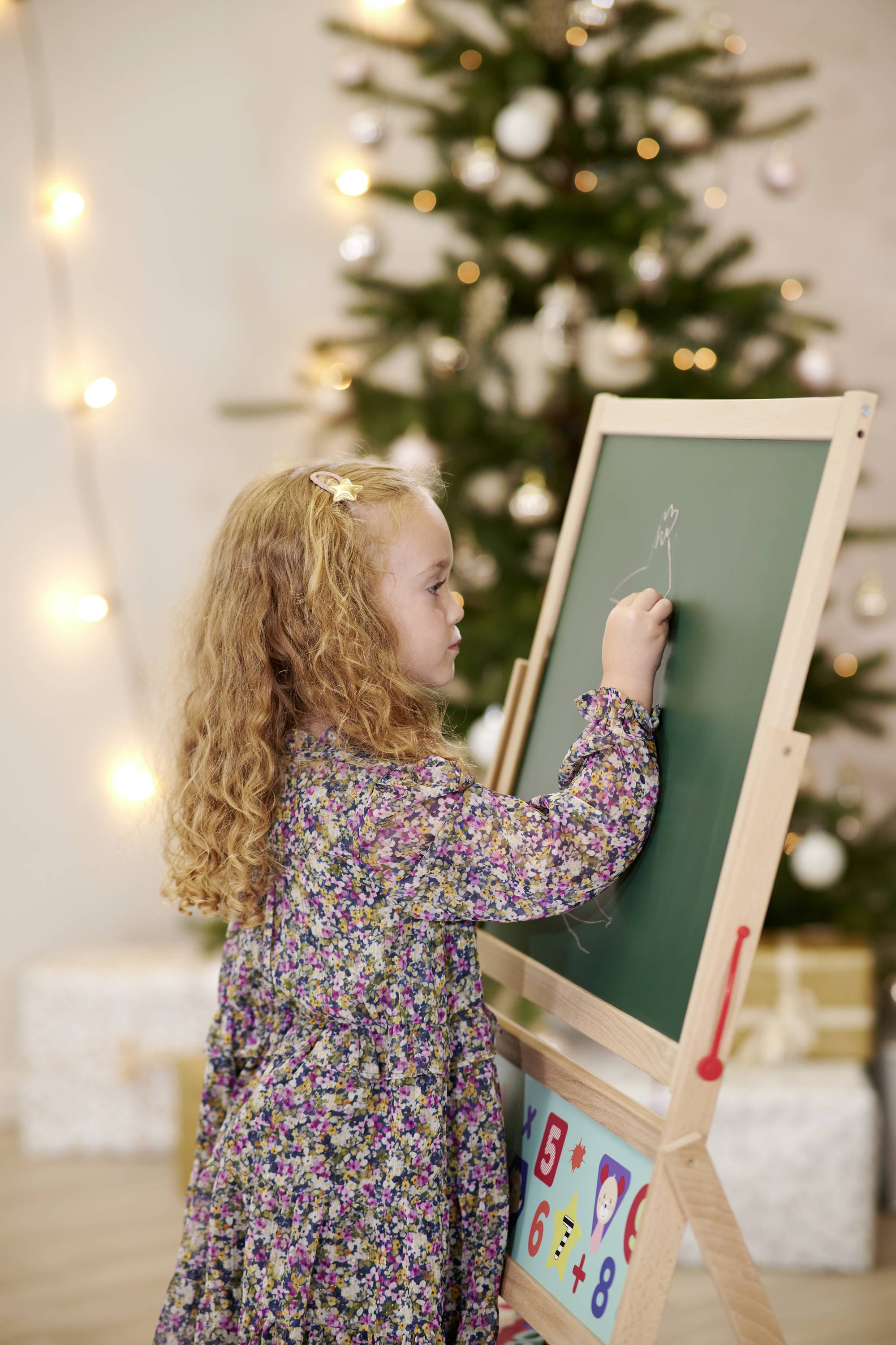 A little girl is drawing on a blackboard in front of a decorated Christmas tree. Lights and wrapped presents are visible in the background.
