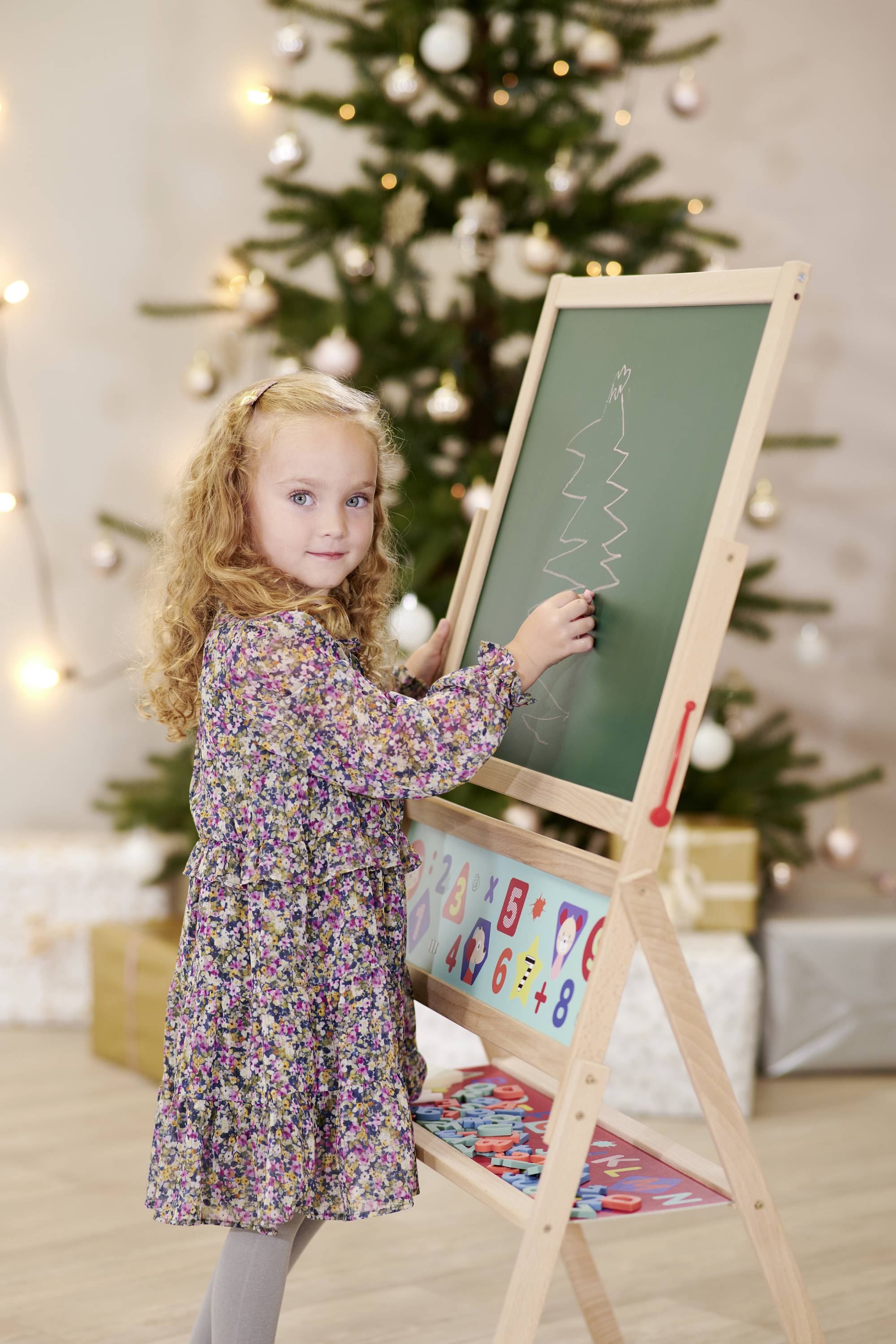 A girl is drawing a Christmas tree on a board. In the background, a decorated Christmas tree stands.