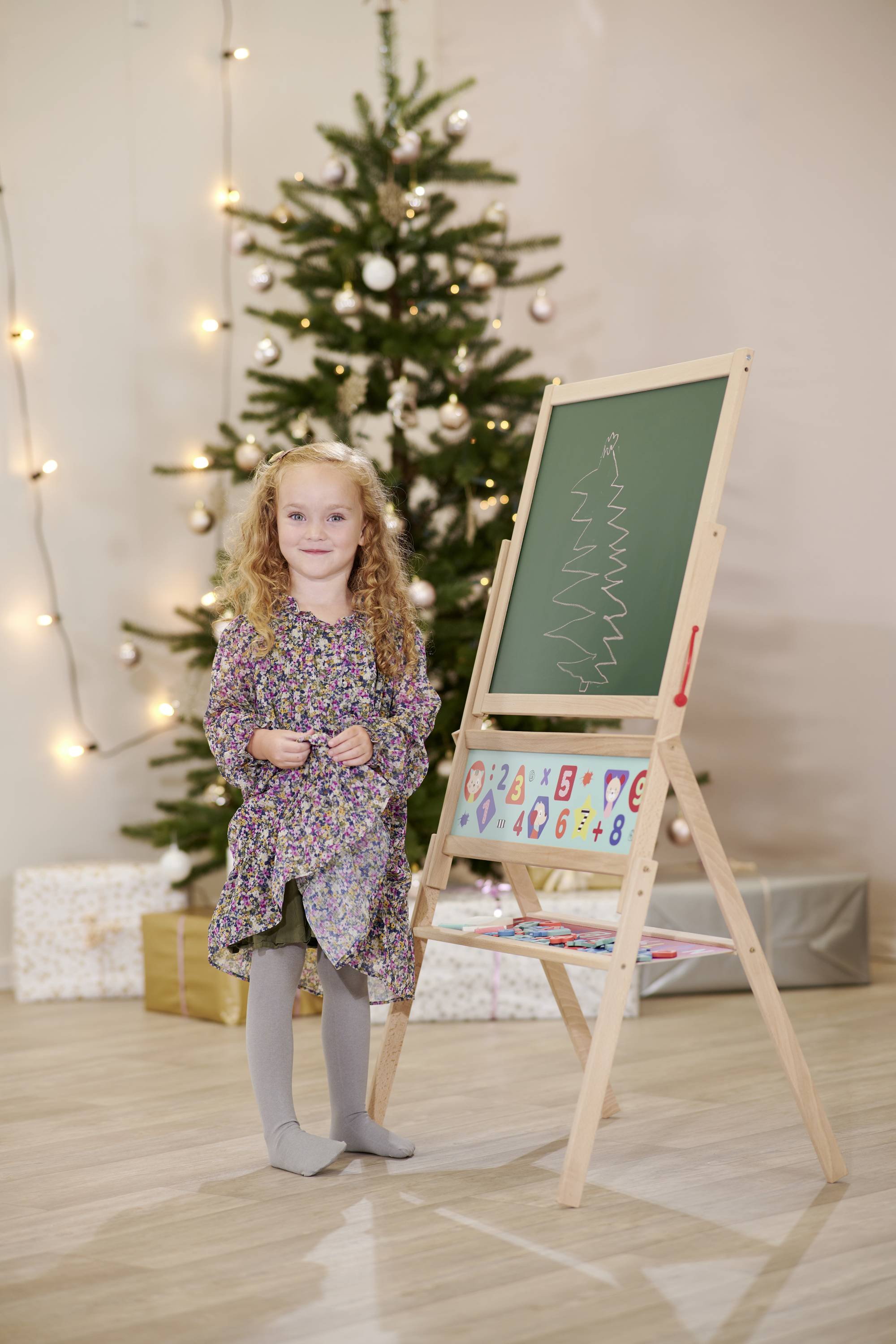 A child is standing next to a blackboard with a drawn pine tree, with a decorated Christmas tree and presents in the background.
