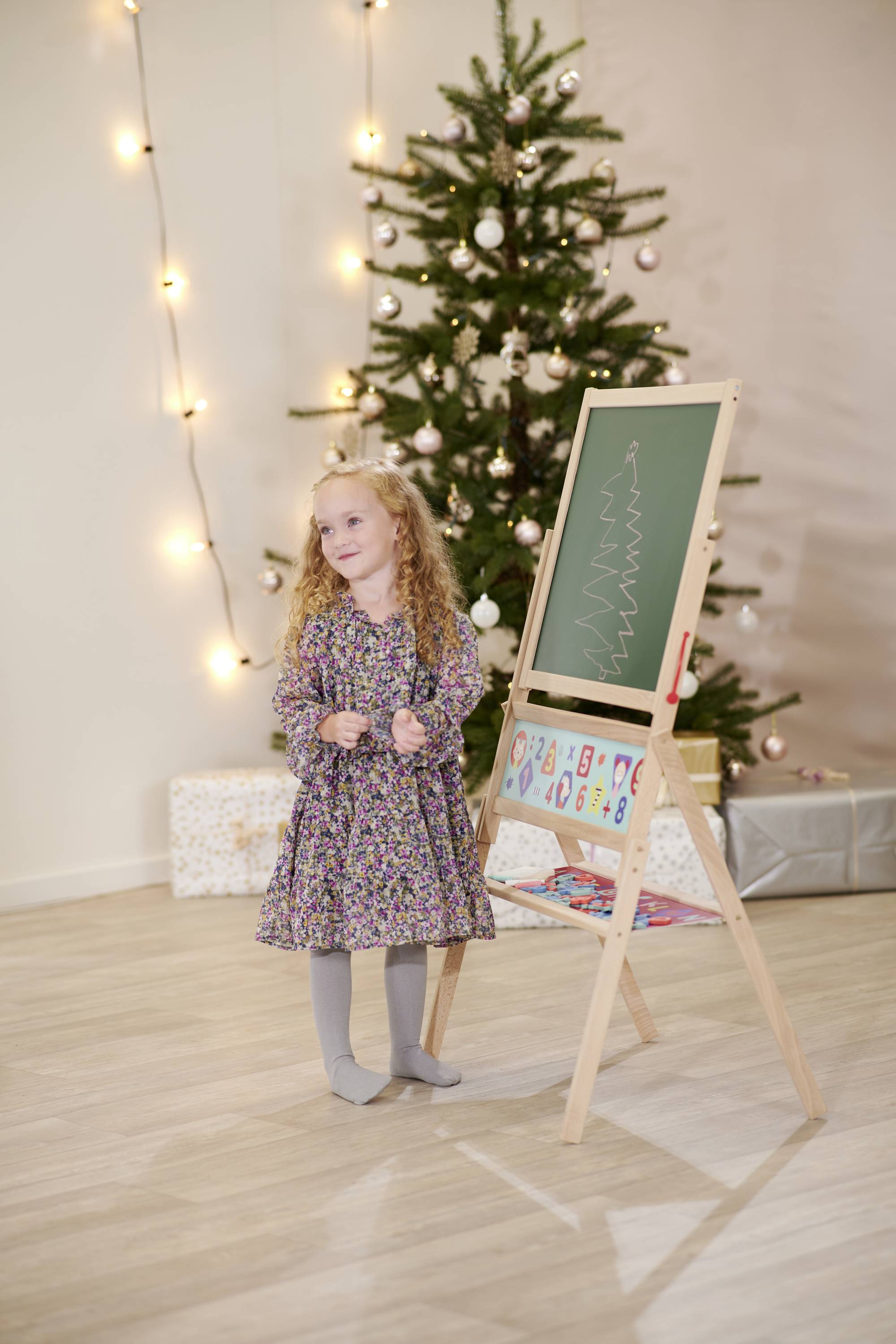A child with curly hair is standing next to a chalkboard drawing, in front of a decorated Christmas tree and presents.
