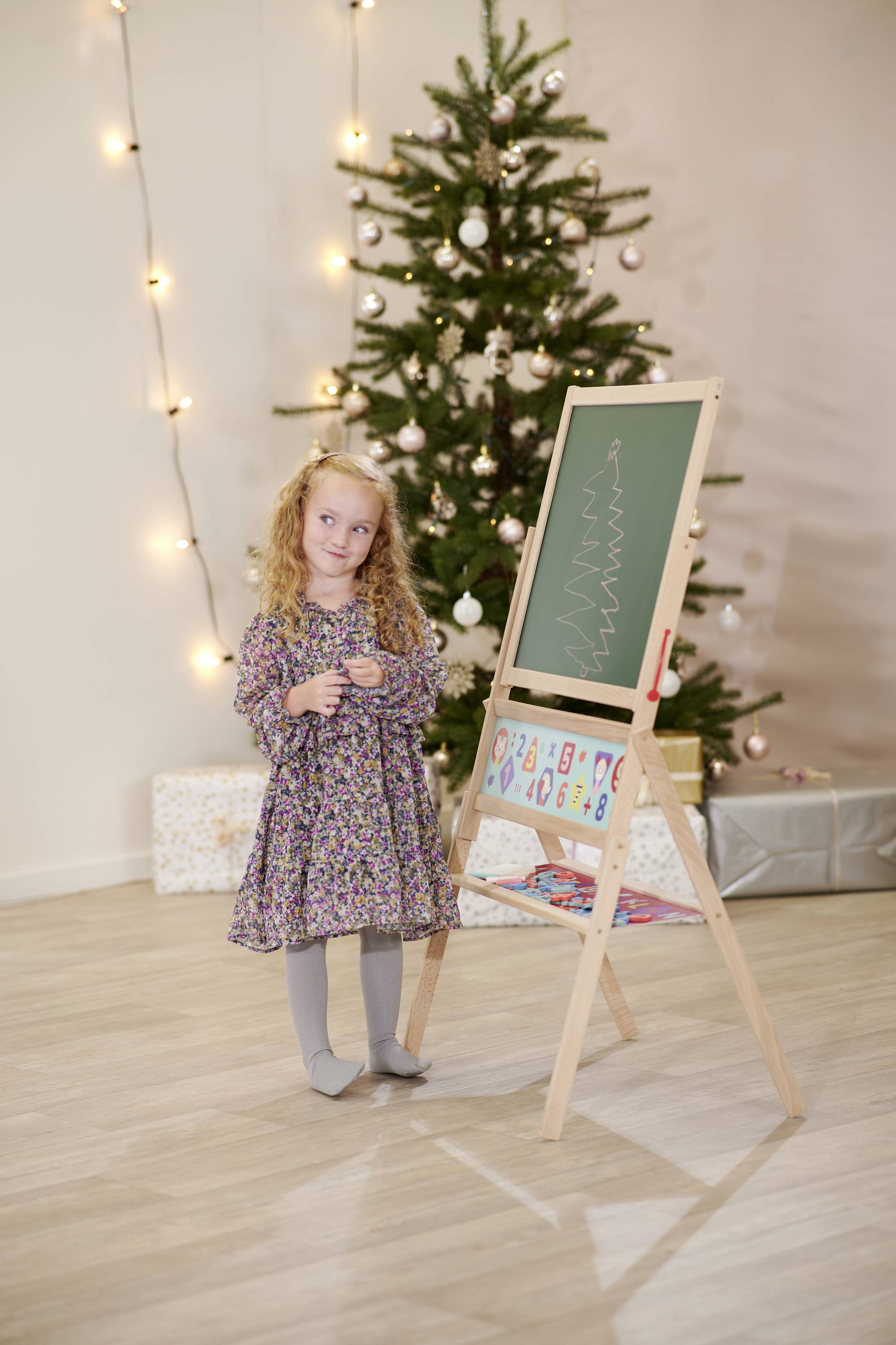 A girl is painting on an easel in front of a Christmas-decorated tree with lights and presents underneath.