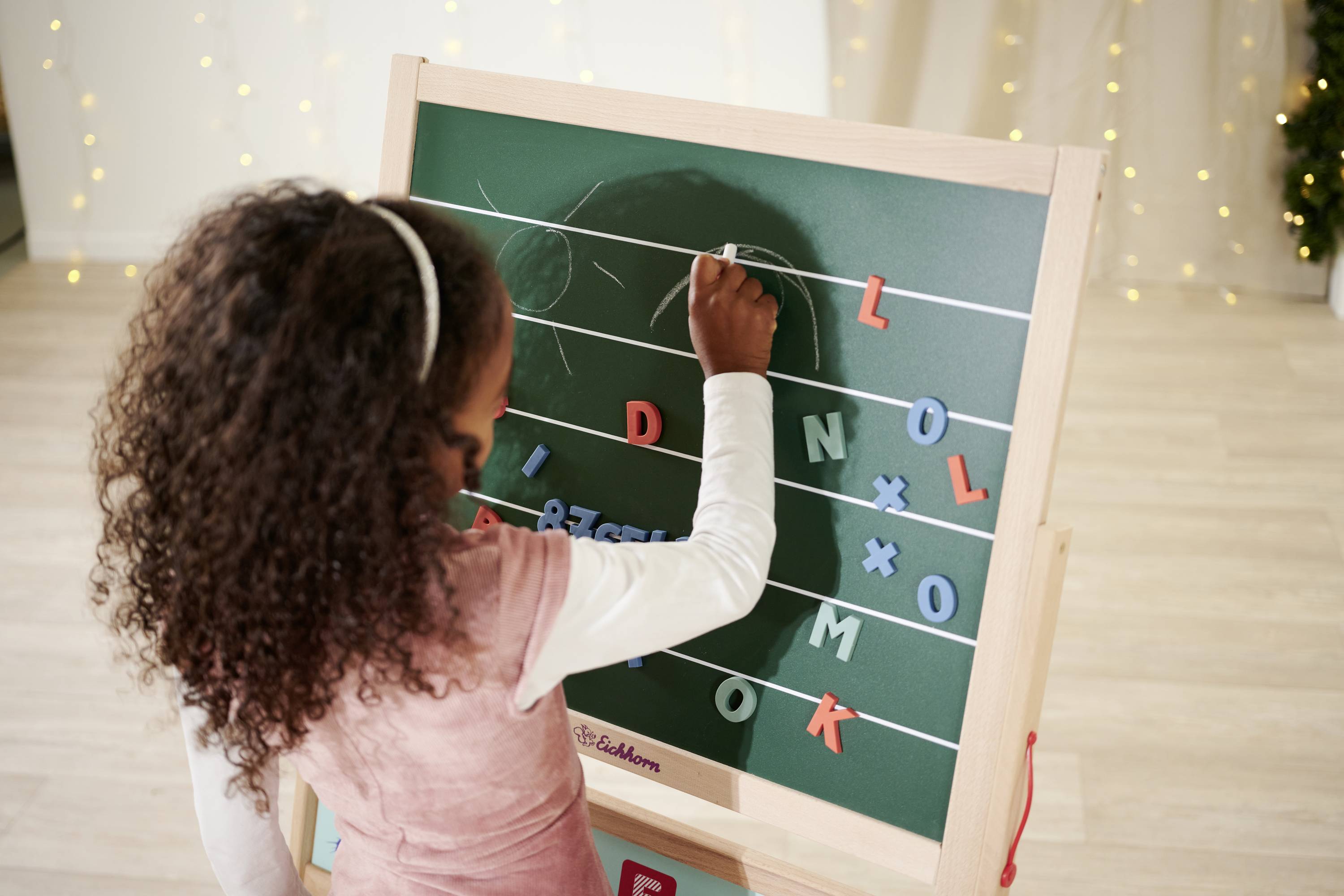 A child is drawing with chalk on a blackboard. Letter magnets are attached to the board. Fairy lights are in the background.