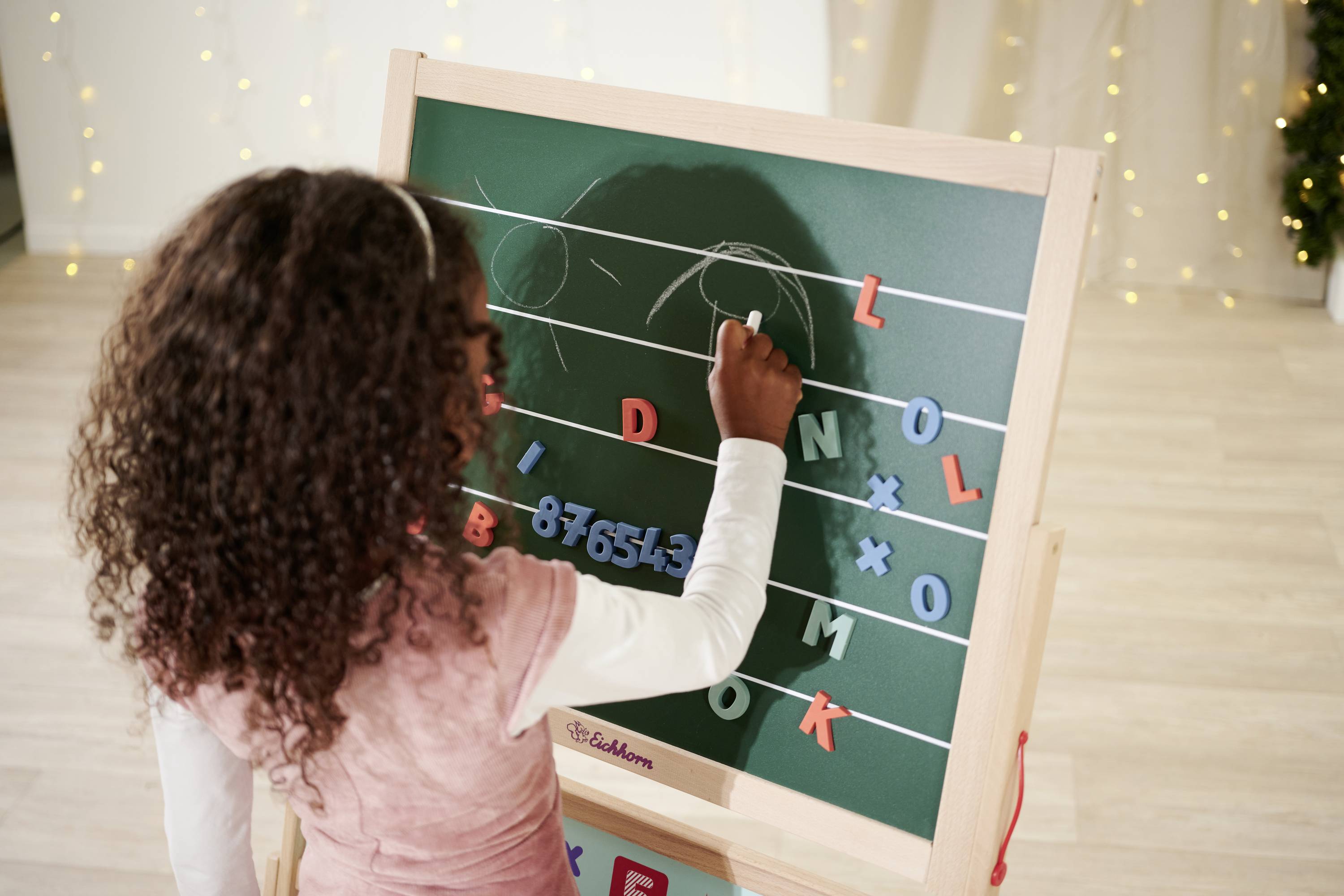 A child writing on a blackboard with letters and numbers, with festive Christmas lights in the background.
