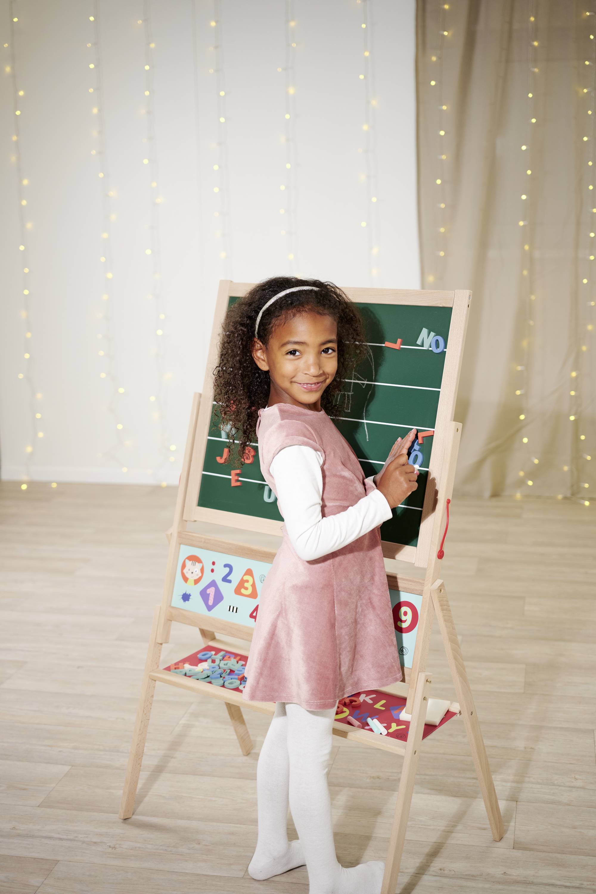 A girl in a pink dress stands smiling at a blackboard and writes with chalk. Background decorated with fairy lights.