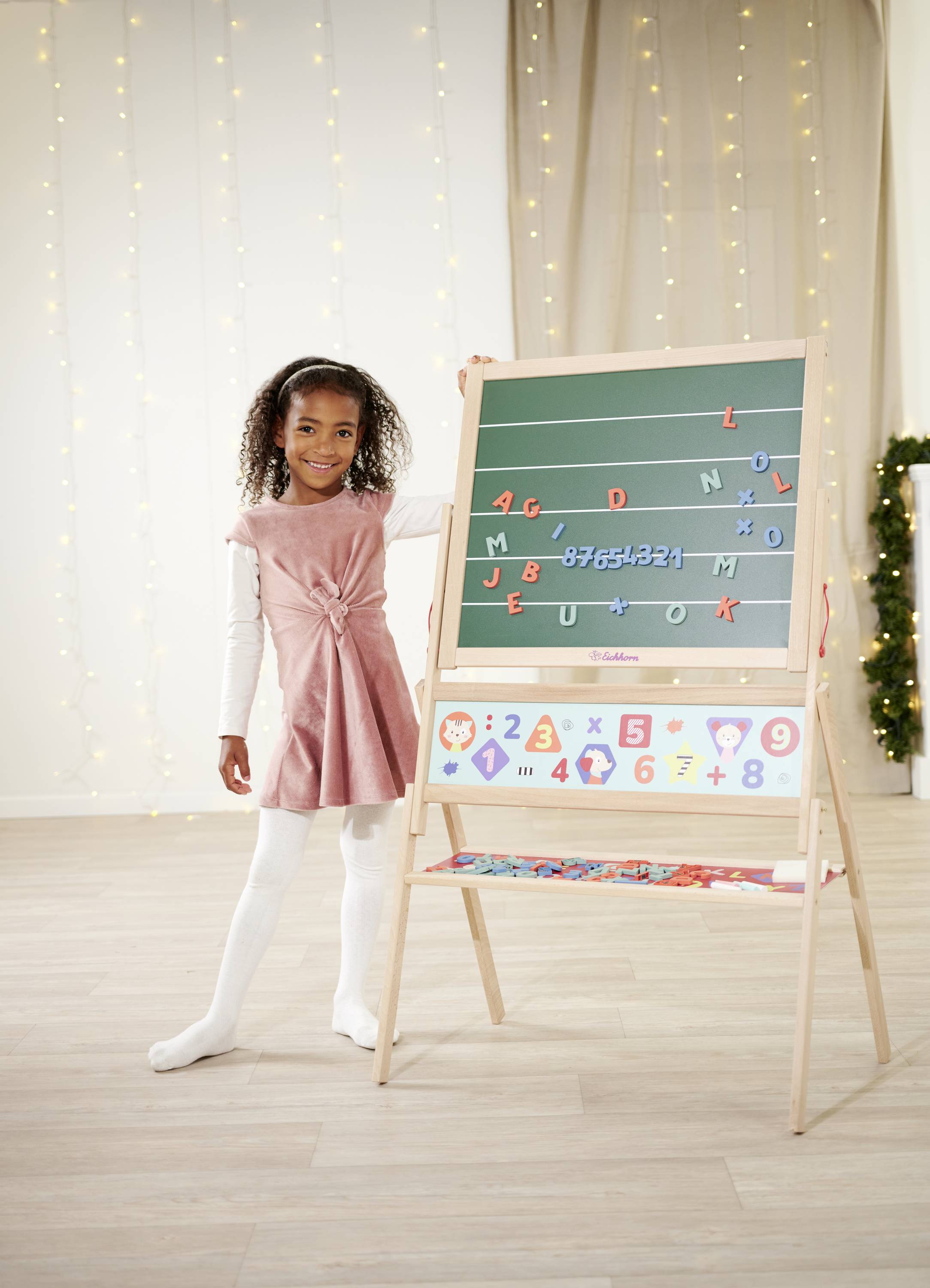 A child smiles beside an easel with a toy alphabet and numbers. Background with lights, festive atmosphere.
