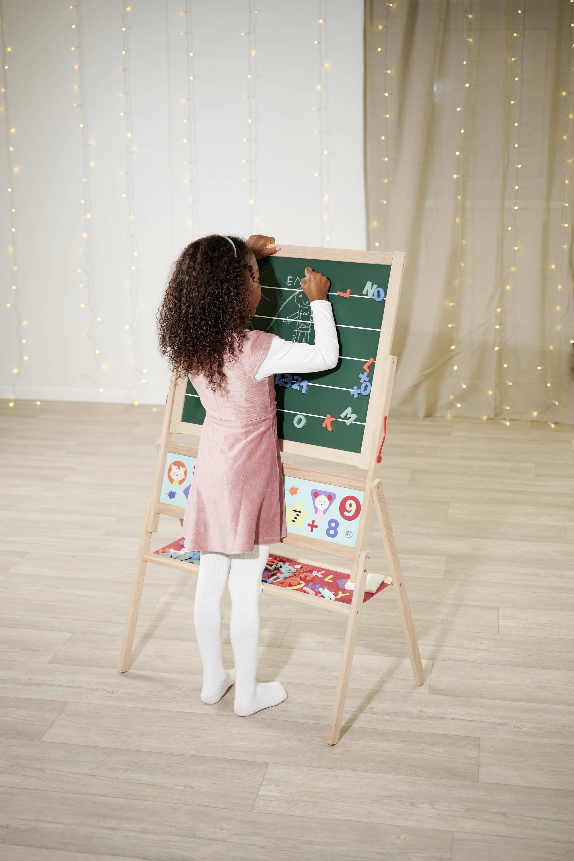 A child is drawing with chalk on a blackboard, with fairy lights hanging in the background.