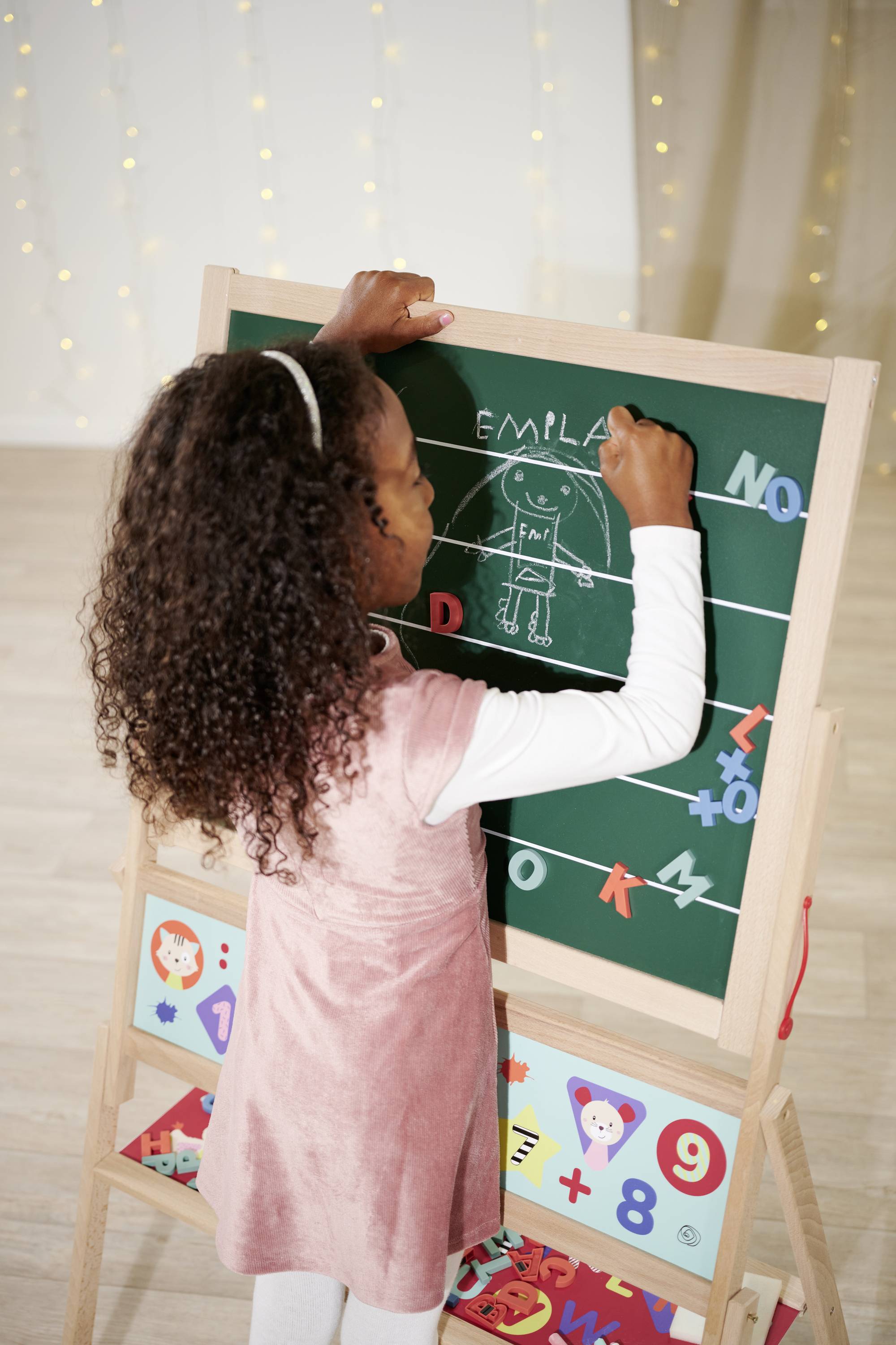A child is drawing with chalk on a blackboard, positioned to the side of the image. The blackboard displays a drawn picture with a name and letters.
