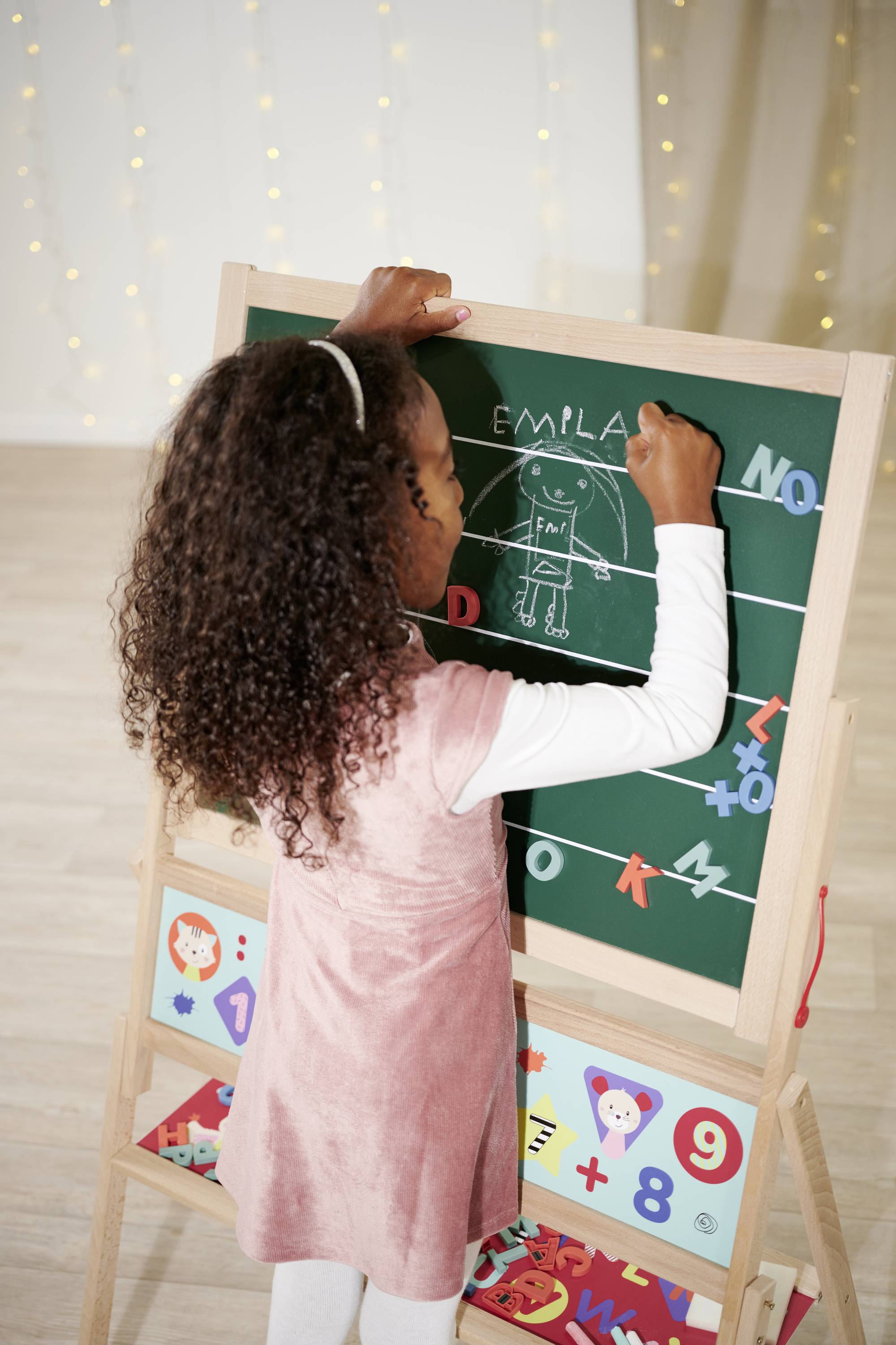 A child is drawing with chalk on a blackboard. They have drawn a figure and are writing some letters above it. Lights are visible in the background.