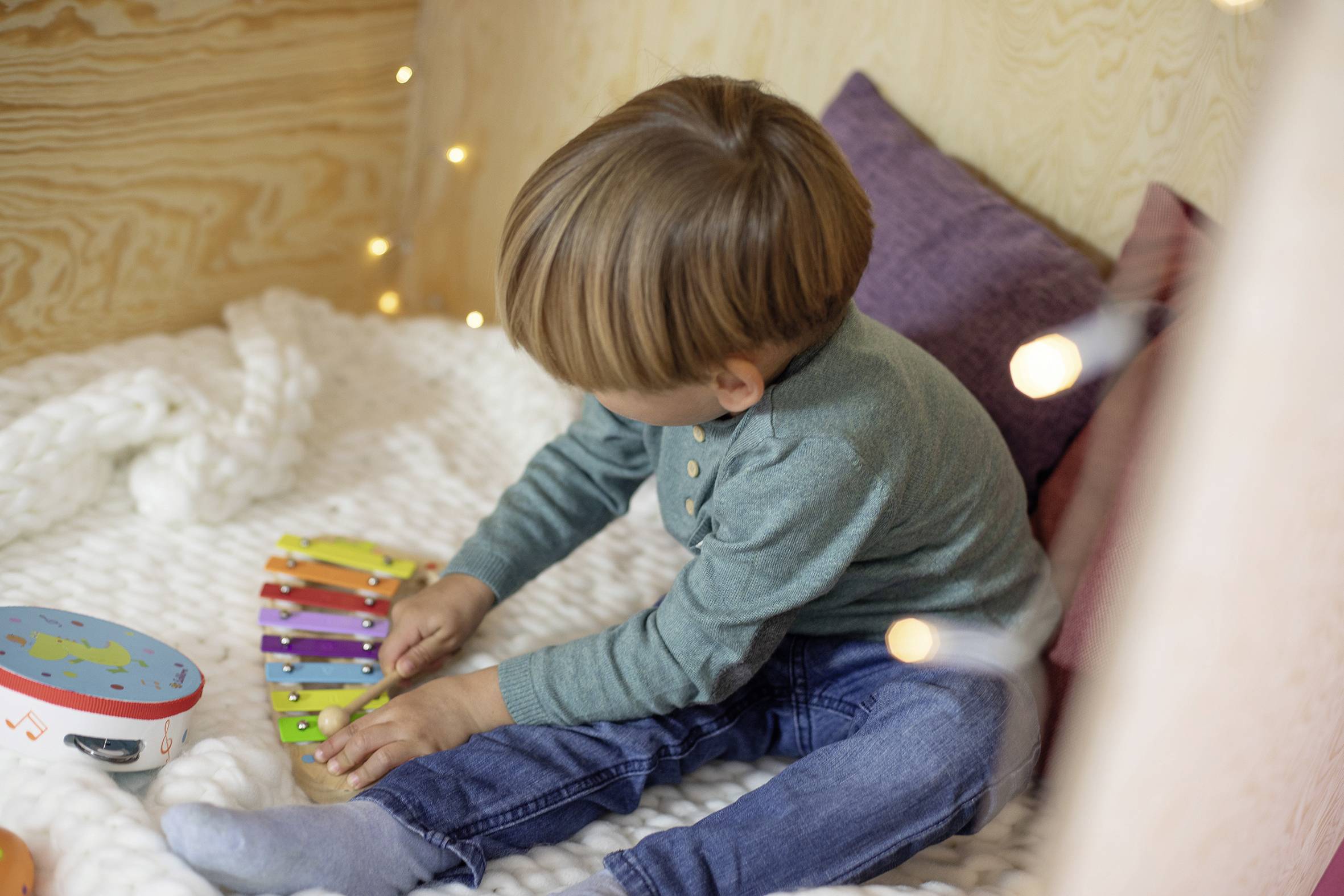 A child is playing on a colourful xylophone. It is sitting cosily on a white cushion in a room with dimmed lighting.