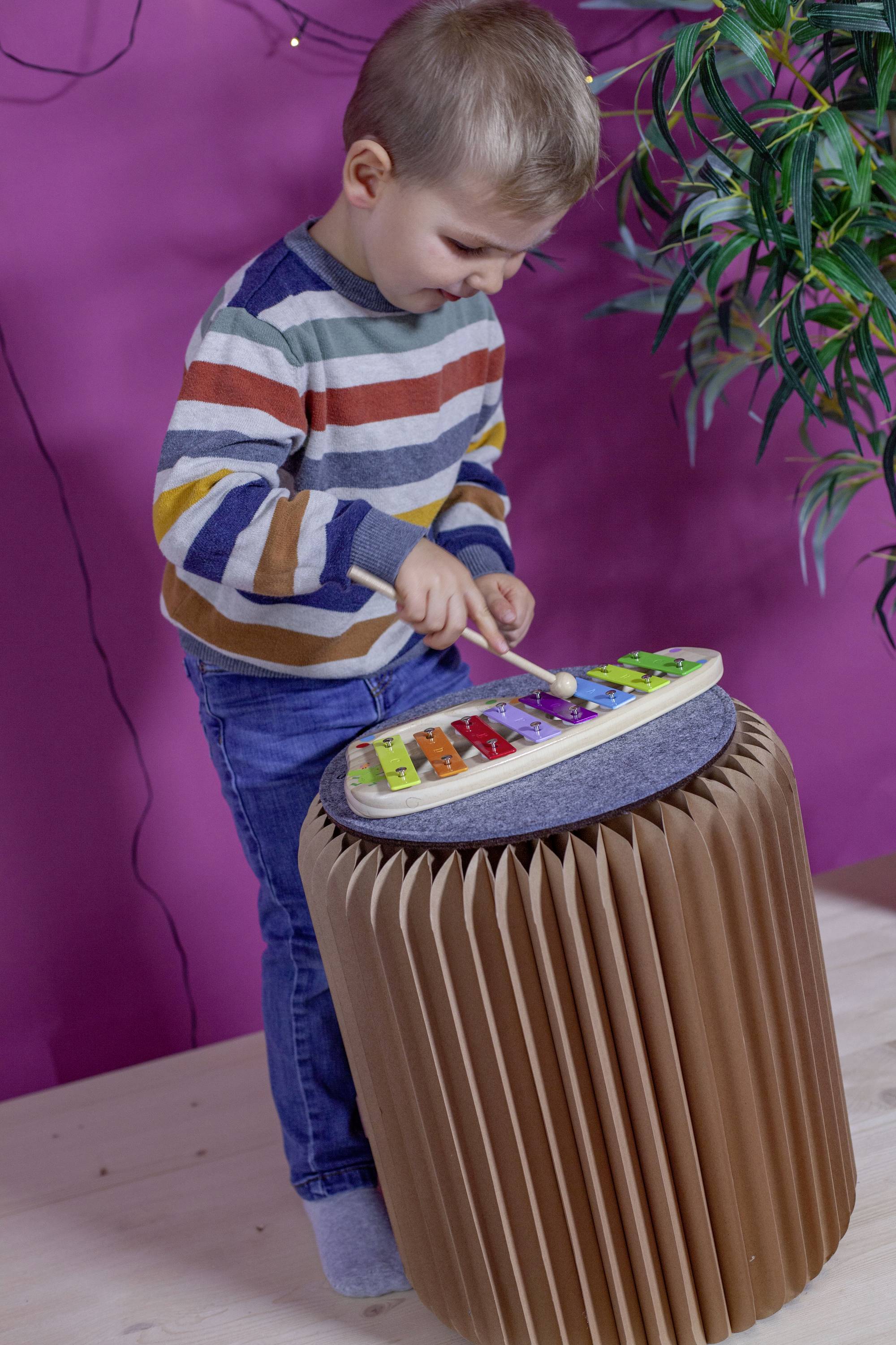 A small boy is playing on a colourful xylophone, which is resting on a brown, paper-like stool. In the background is a lilac wall.