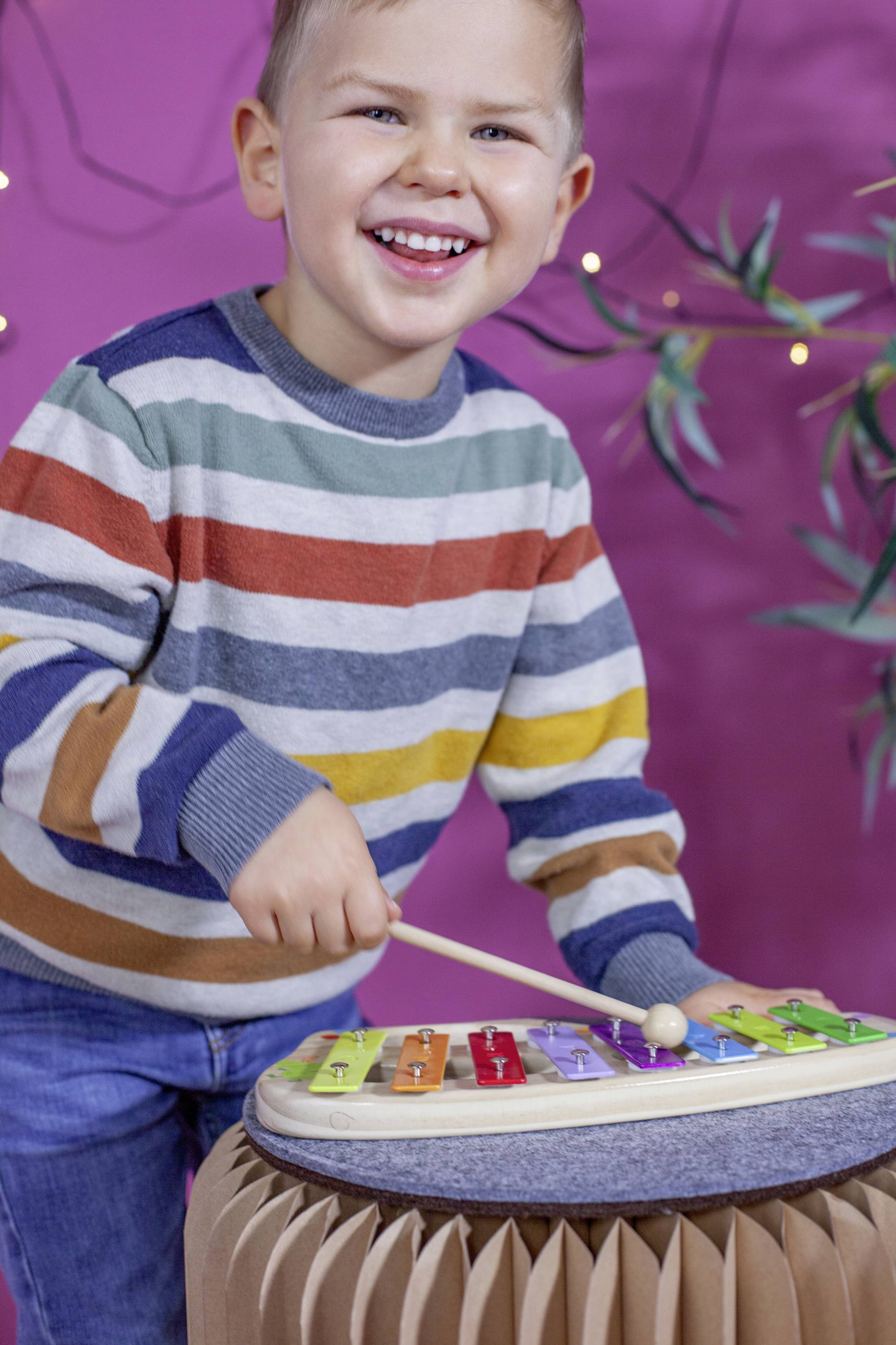 A child in a striped jumper plays cheerfully on a colourful xylophone in front of a pink background with fairy lights.