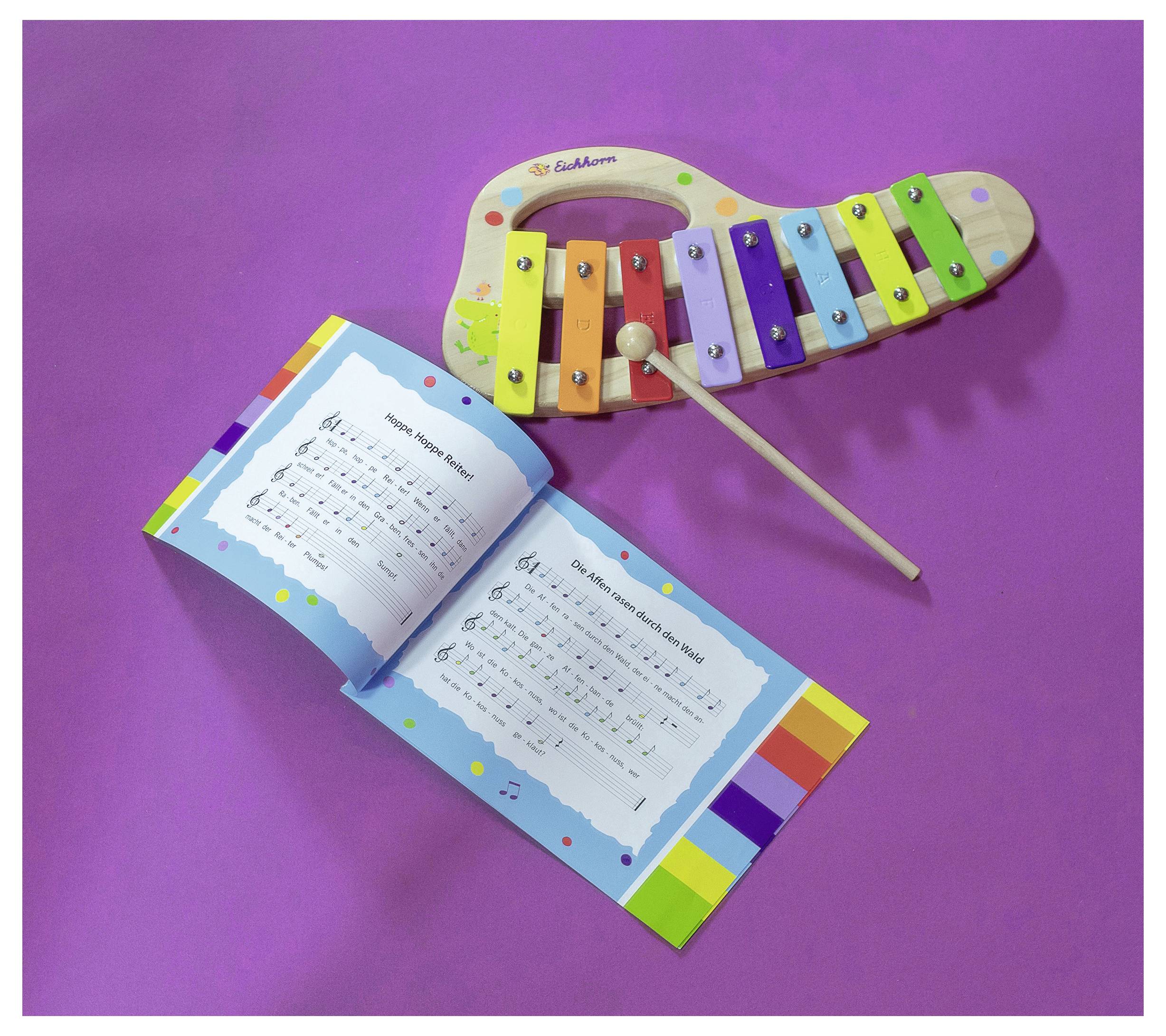 A colourful xylophone and an open children's songbook are lying on a lilac background. The book displays musical notes and song lyrics.