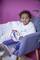 A small child sits in a grey armchair against a pink background. They are holding a tambourine and wearing white clothing and jeans.