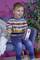 A smiling child wearing a striped jumper sits on a chair and holds a toy. Plants are visible in the background.