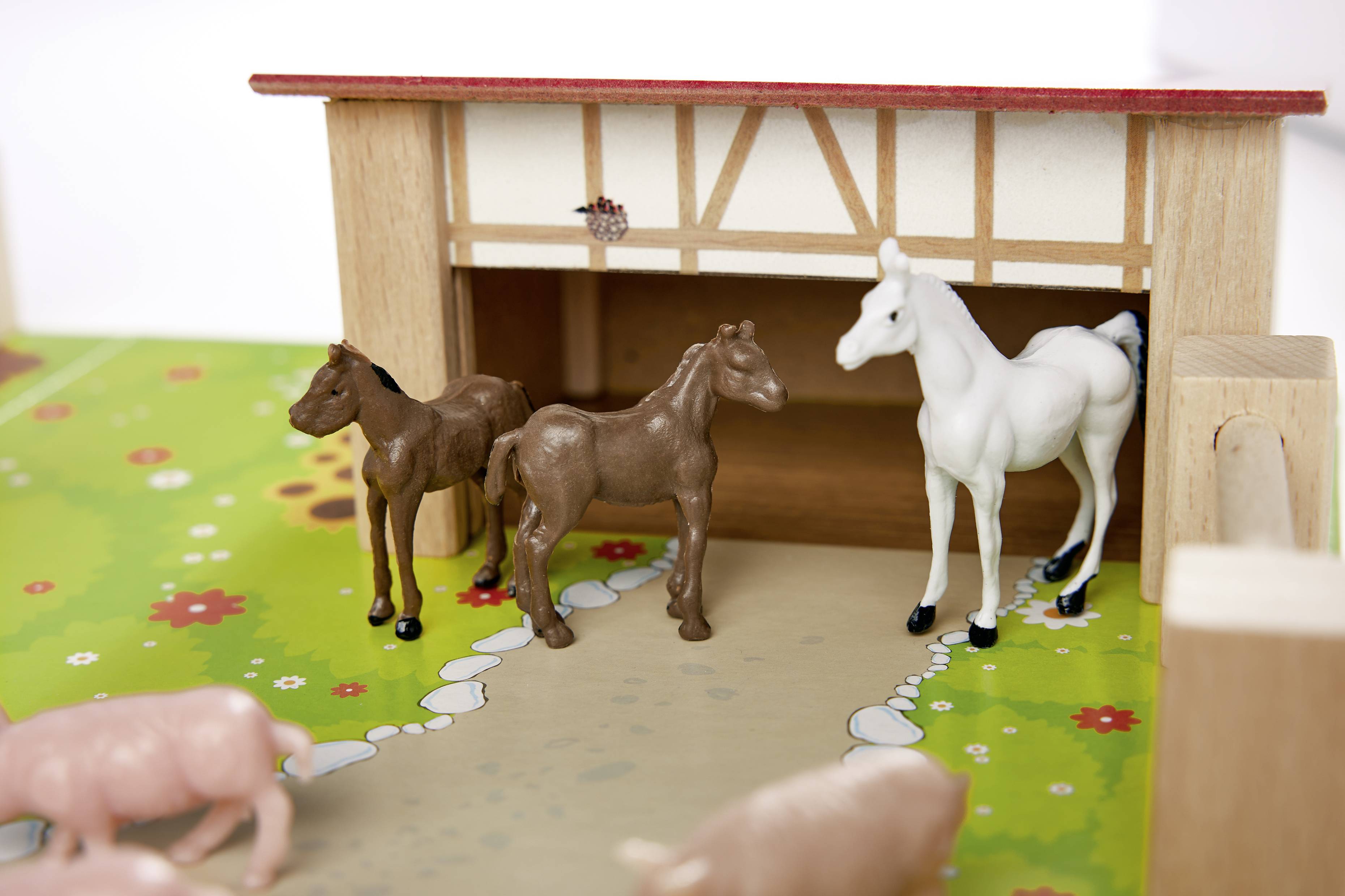 'Toy horses in a stable next to pigs on a colourful play mat. The stable is made of wood with a red roof, with floral patterns in the background.'