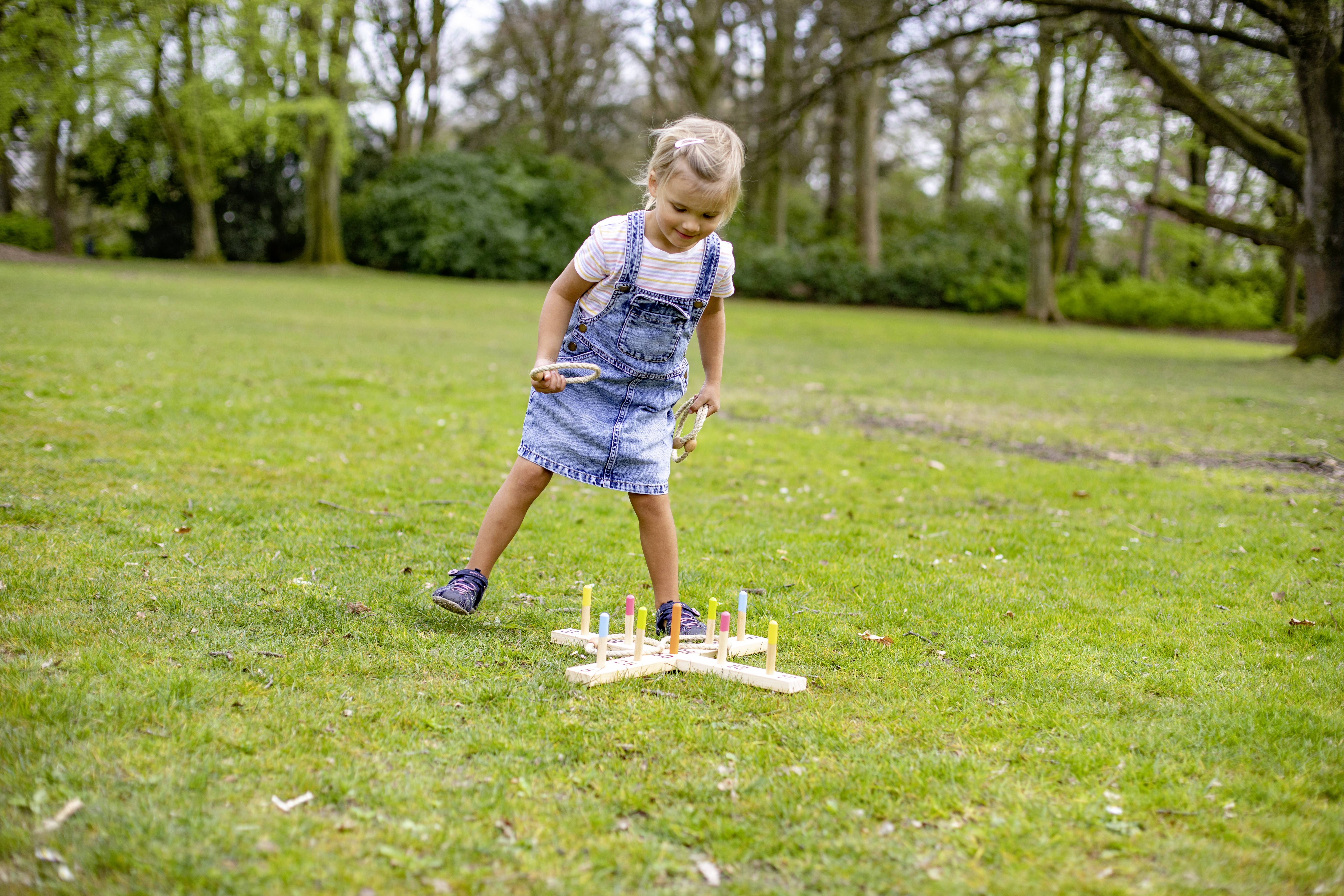 A child is playing outdoors with a ring toss game on grass-covered ground. Surrounded by trees under a sunny sky.
