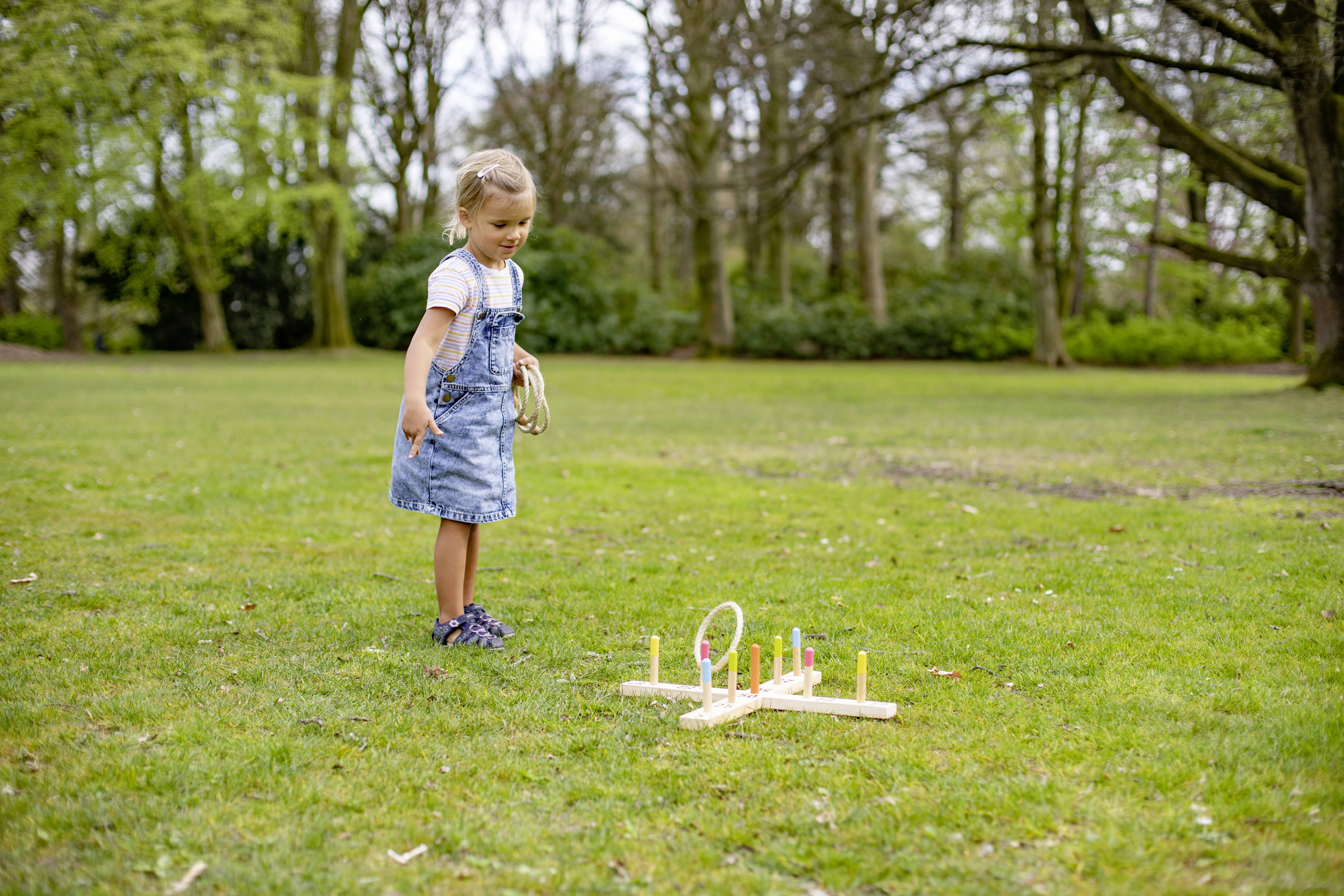 A small girl throws a ring onto a ring toss game in the park. She is wearing a denim dress and is focused on the game.