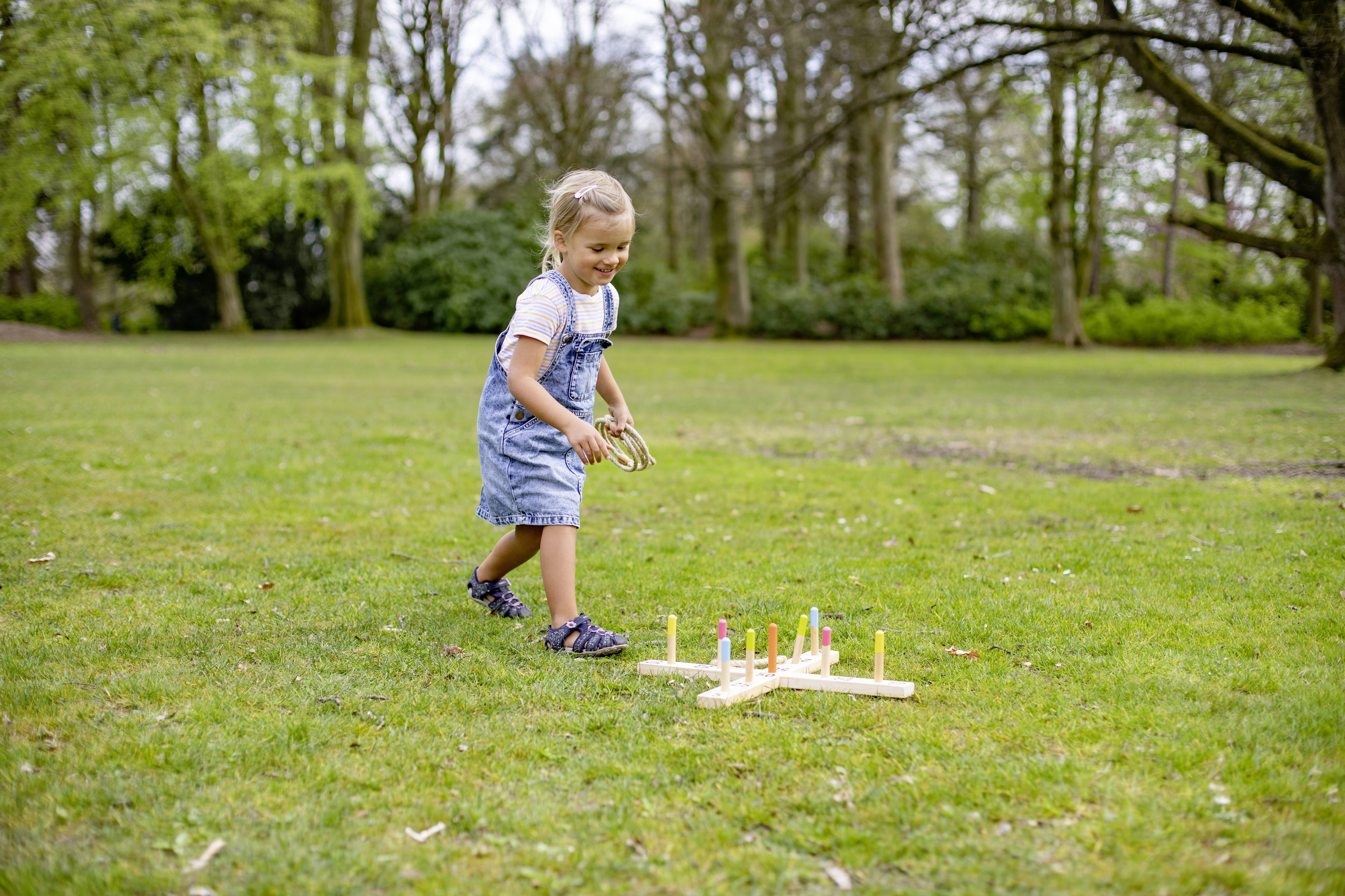 A child cheerfully throws rings onto a wooden target game on a lawn. Trees and green surroundings in the background.